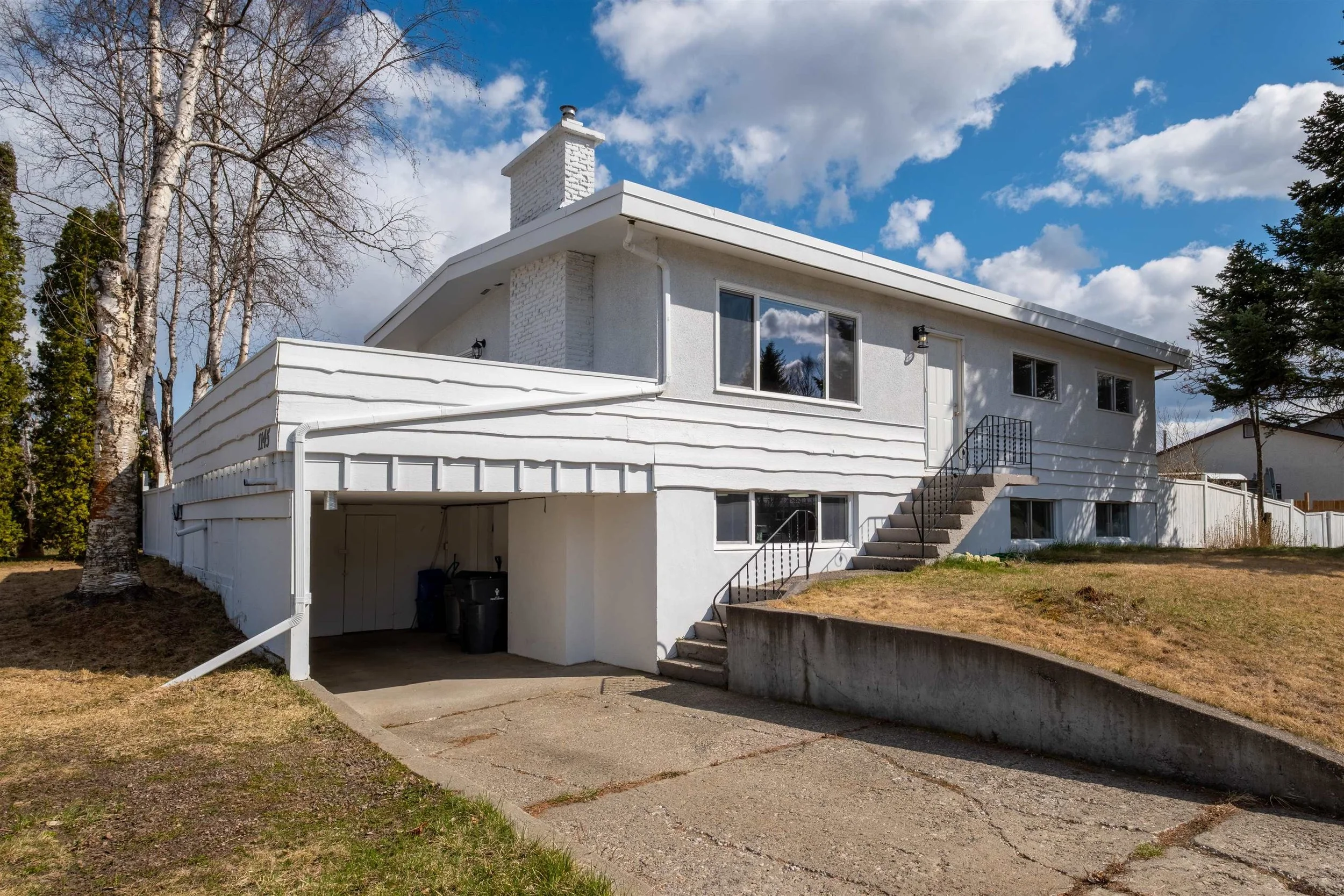 A white two-story house with a sloped roof, set against a partly cloudy sky. The house has large windows, a brick chimney, and external stairs leading to the door. A driveway and garage are visible.