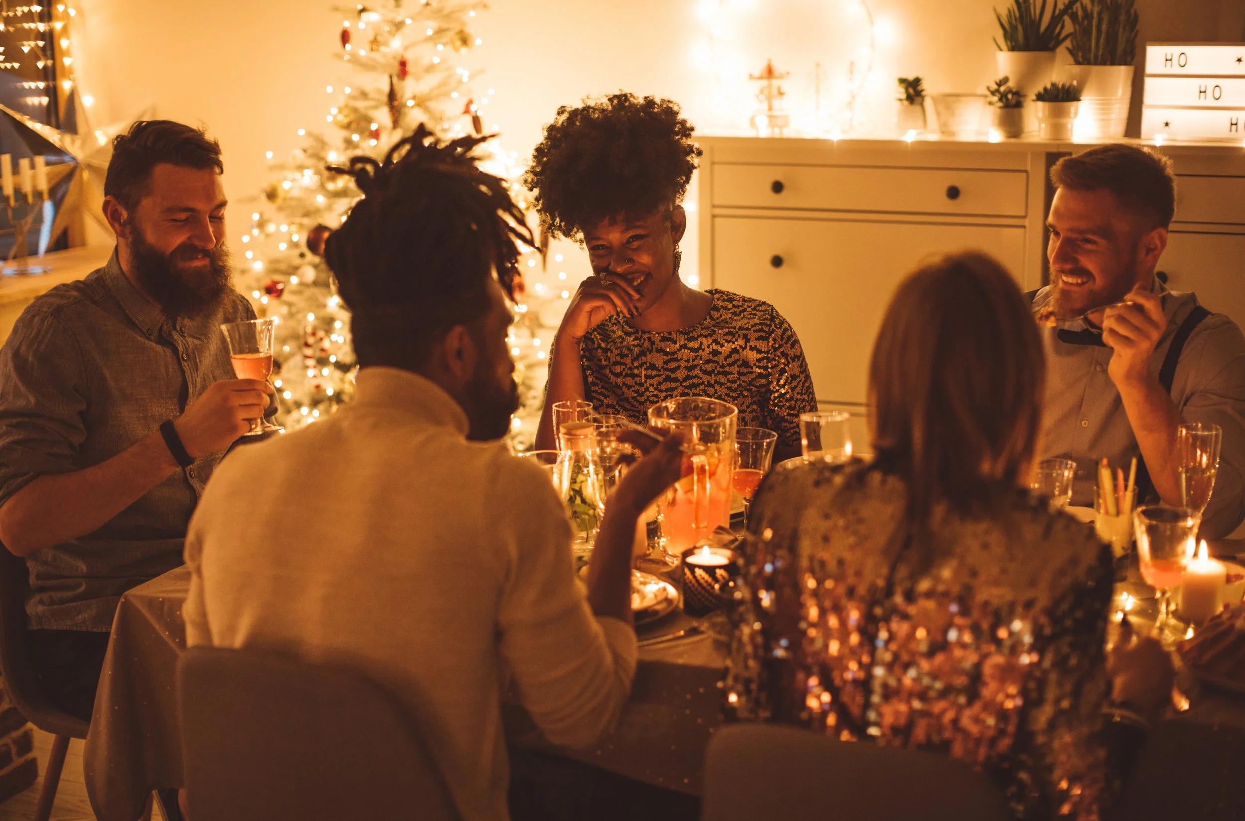 Group of six friends gathered around a dinner table, smiling and laughing, with a decorated Christmas tree and warm lights in the background.