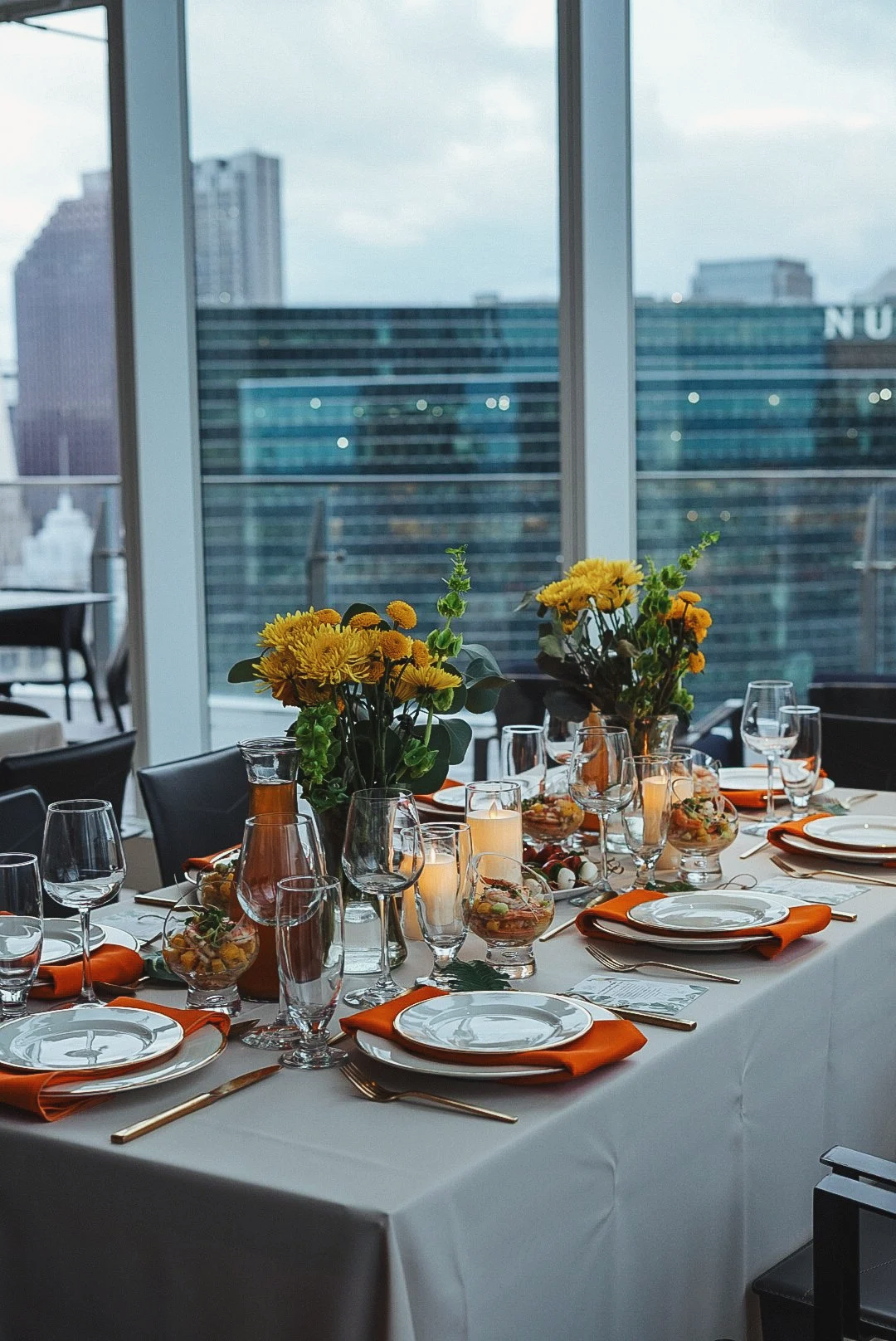 A dining table set for a celebration with white tablecloth, orange napkins, multiple glasses, and floral centerpieces with yellow flowers in vases.
