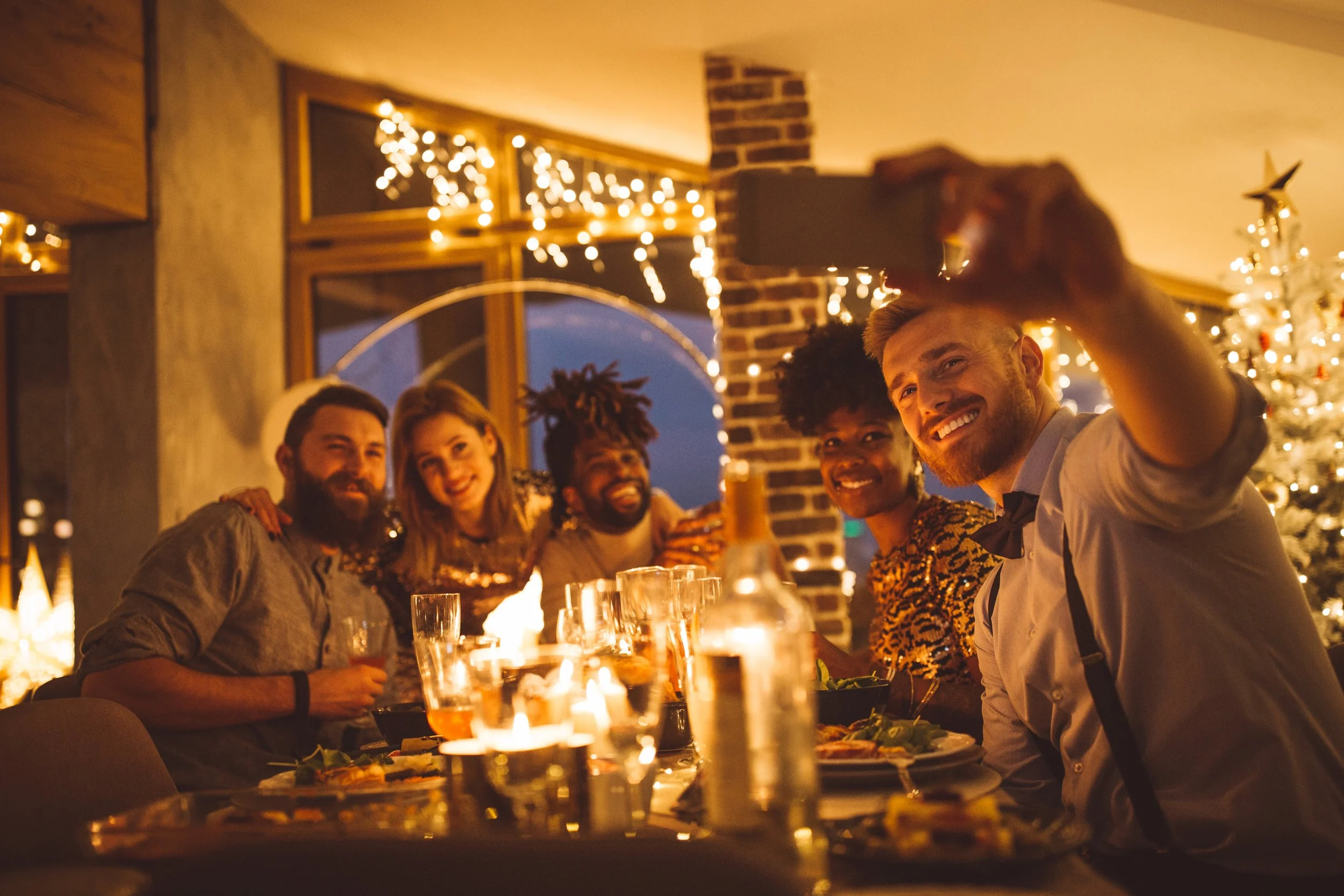 Group of friends enjoying a dinner party with Christmas decorations, including a Christmas tree and string lights, in a cozy indoor setting during evening.