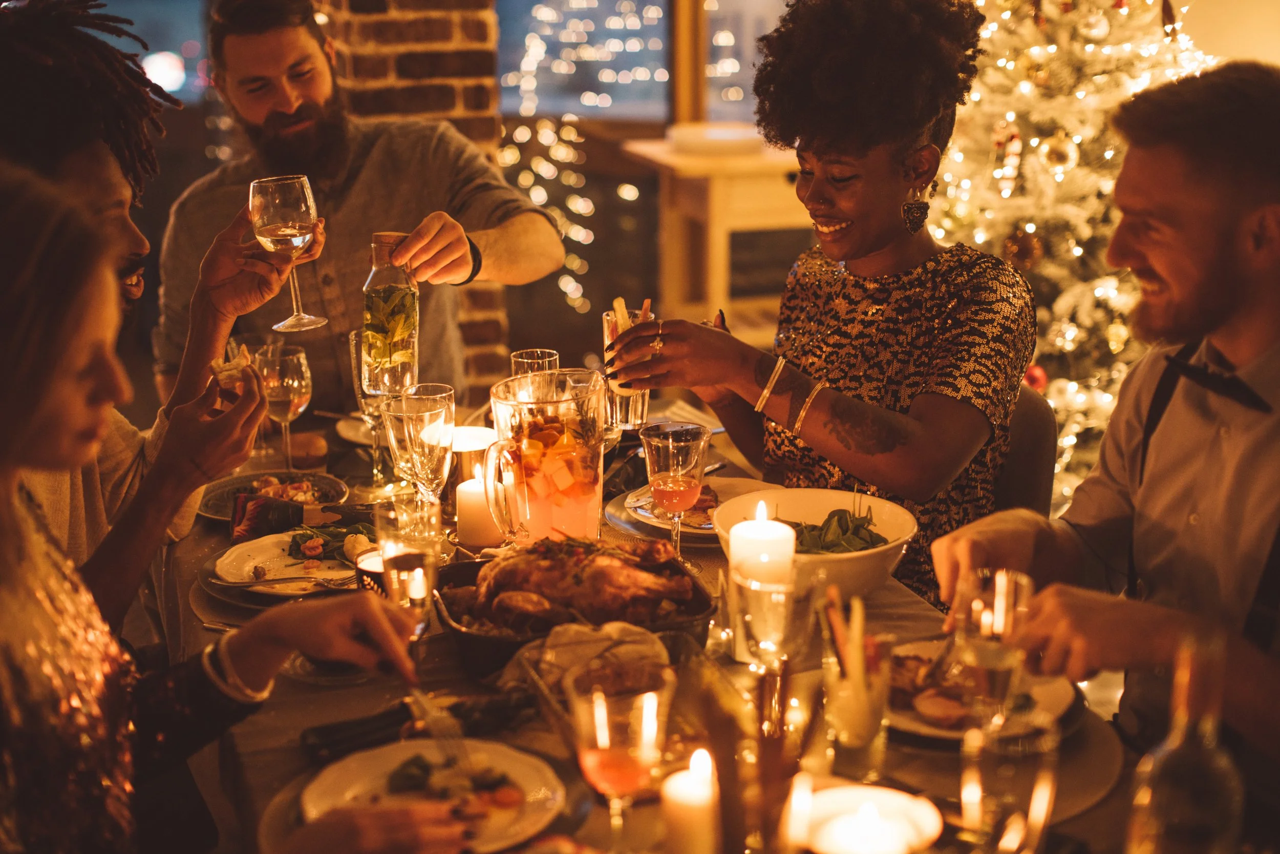 A diverse group of friends celebrating Christmas dinner around a table decorated with candles and a Christmas tree in the background, enjoying food and drinks.