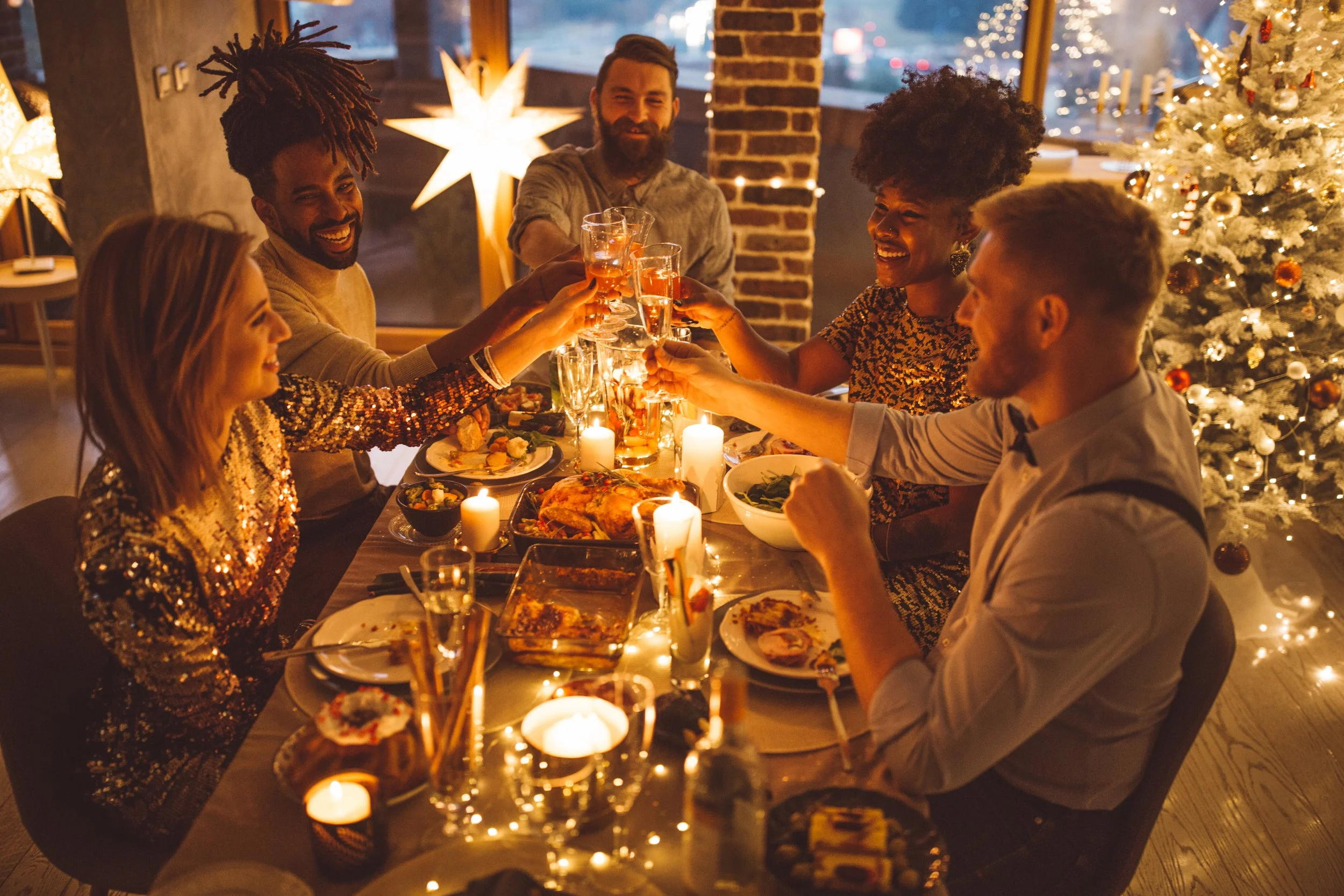 A group of six diverse people celebrating a Christmas dinner, raising glasses in a toast, with a decorated Christmas tree and candles on the table.
