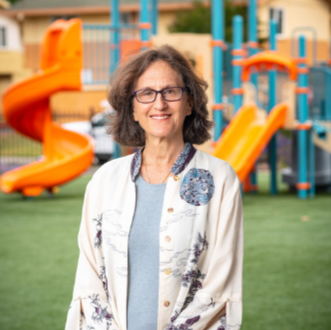 Bina Lefkovitz. A smiling woman with curly hair and glasses standing outdoors in front of colorful playground equipment.