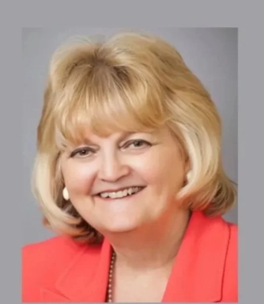 Pam Costa. A smiling middle-aged woman with blonde hair, wearing a coral blazer and pearl earrings, against a gray background.