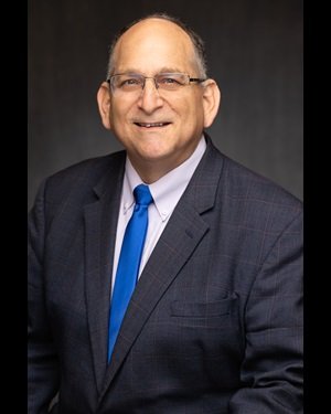 Gregg Fishman. A middle-aged man wearing glasses, a dark suit, white shirt, and blue tie, smiling against a dark background.