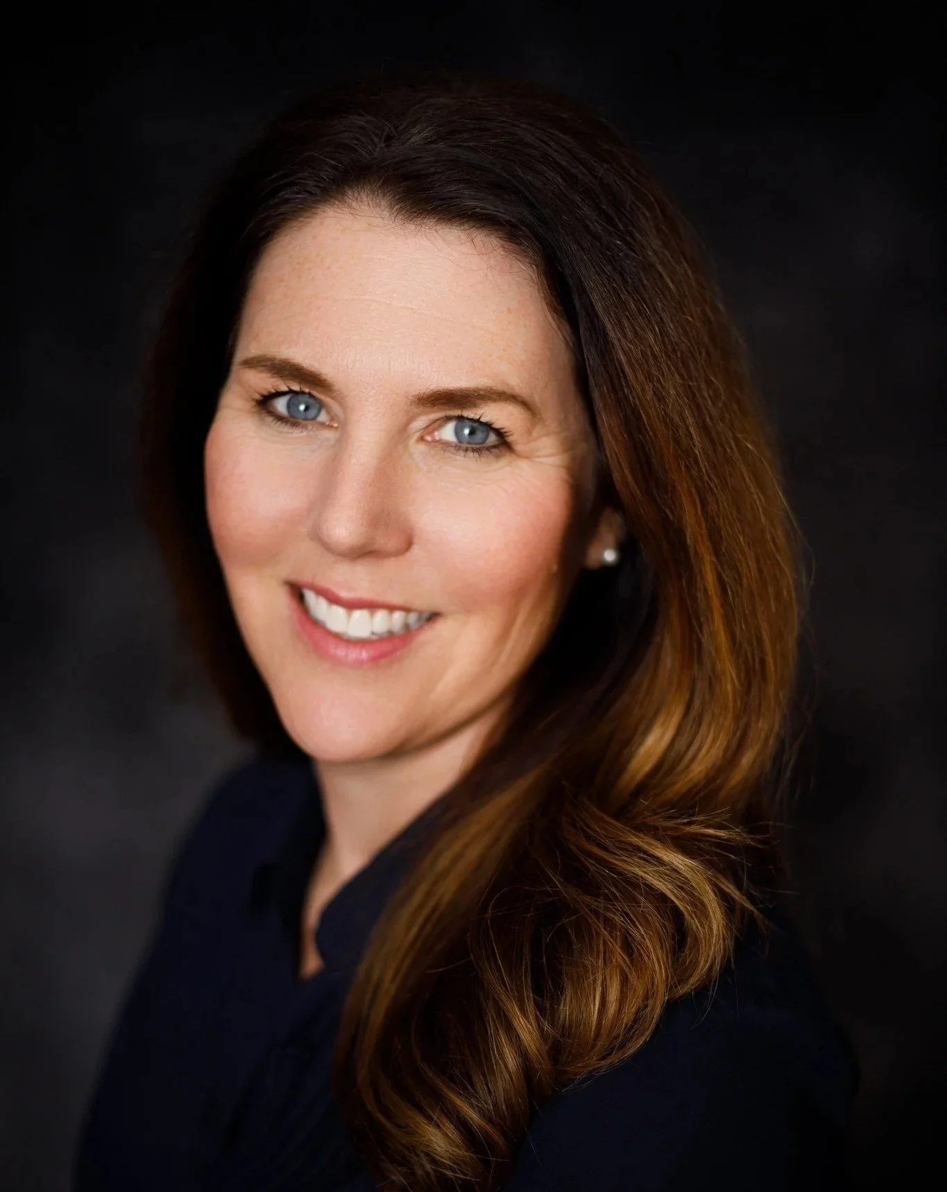 Sarah Kirby-Gonzalez. A woman with long, wavy brown hair and blue eyes smiling, wearing a dark top, posed against a dark background.