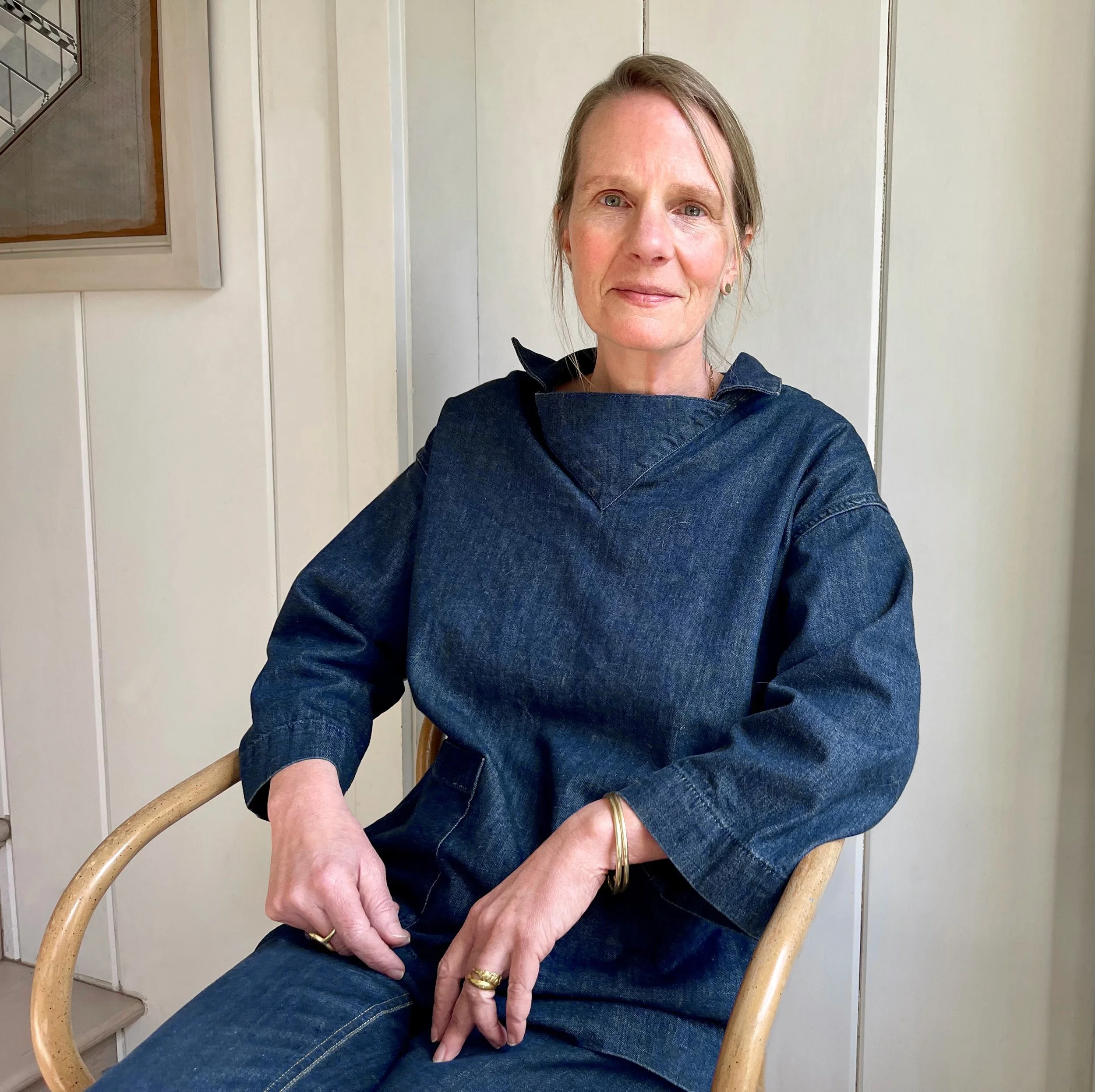 Anne Johnson sitting on a wooden chair, wearing a denim shirt and jeans, with light-colored hair, sitting indoors against white paneled walls.