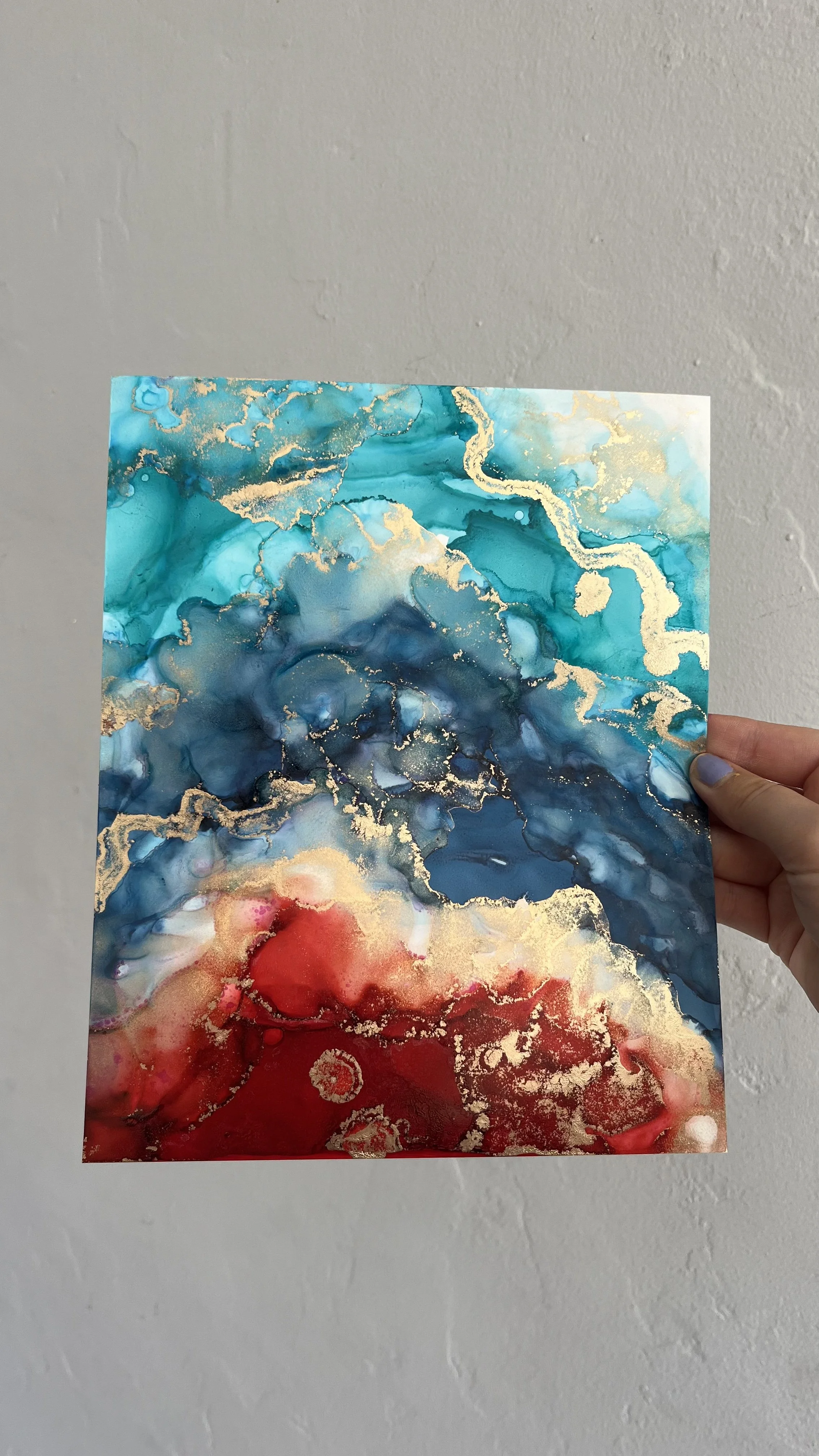 A person holding an abstract watercolor painting with swirling shades of blue, red, and gold accents, against a plain wall background. This is an aura painting and it comes with an aura reading. An aura reader & aura artist made this.