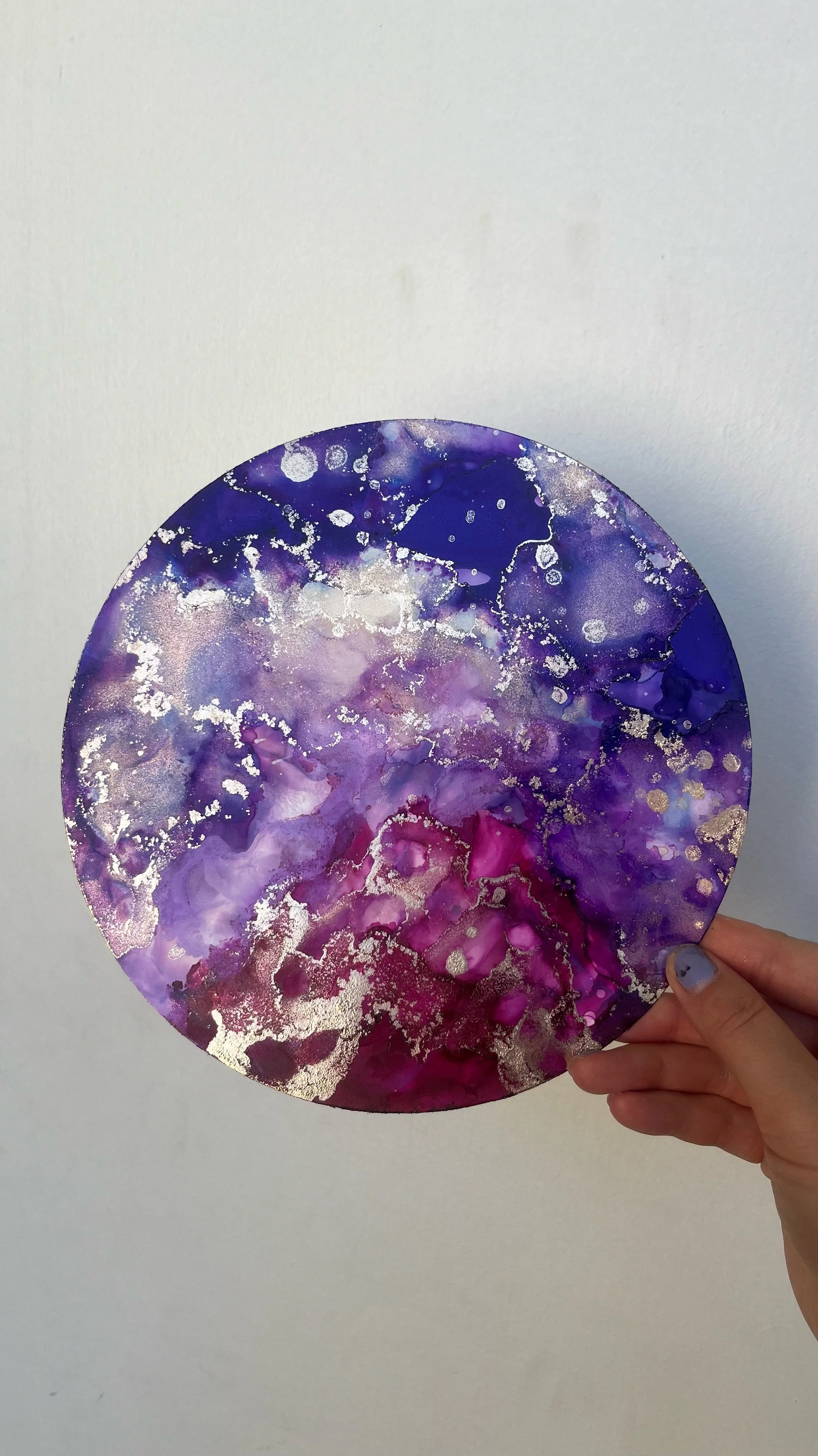 A hand holding a circular, galaxy-inspired abstract art piece with purple, pink, and blue hues and metallic gold accents. This is an aura painting and it comes with an aura reading. An aura reader & aura artist made this.
