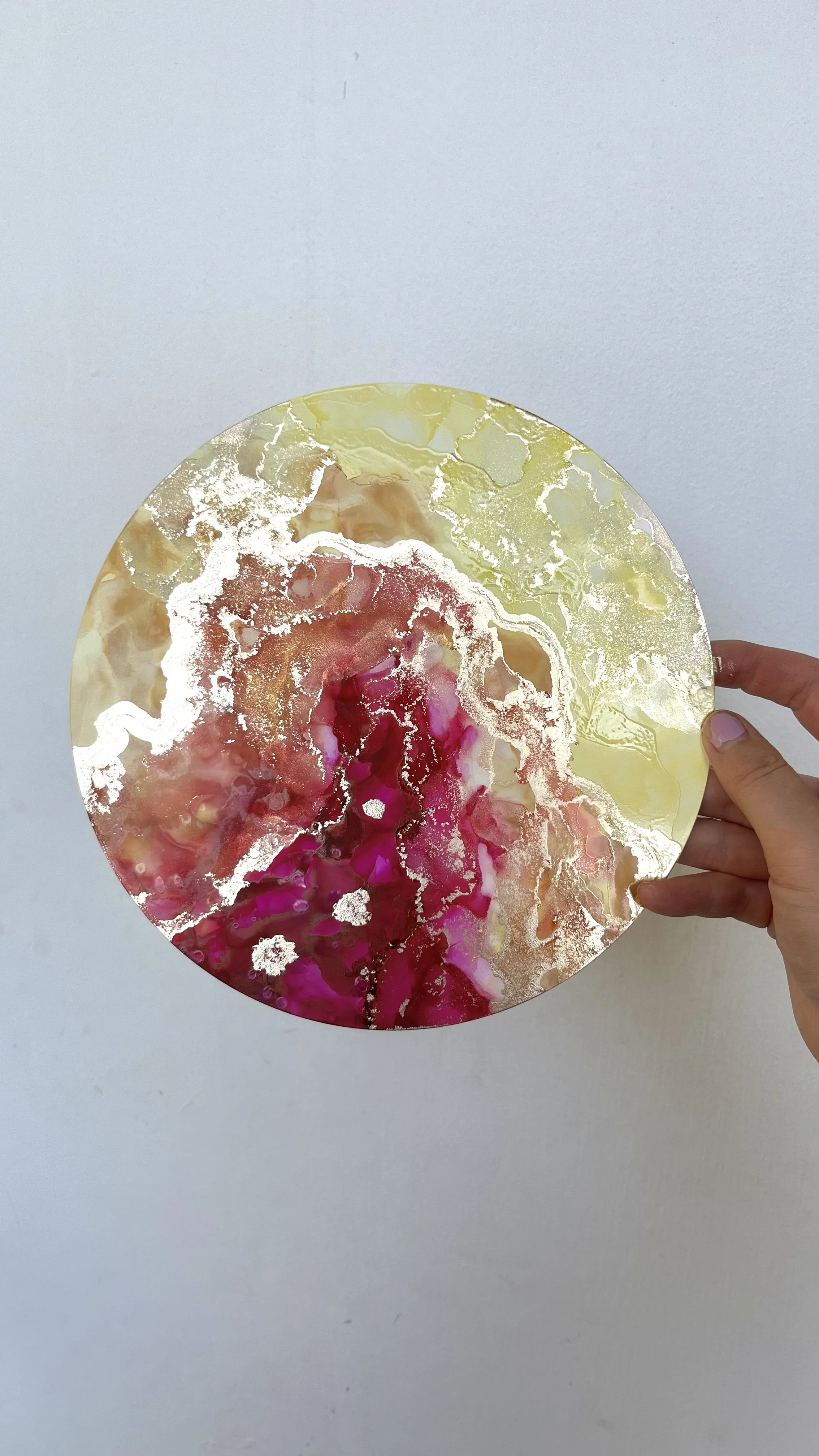 A round, colorful abstract art piece with shades of pink, yellow, and white, held by a person's hand against a plain white wall. This is an aura painting and it comes with an aura reading. An aura reader & aura artist made this.