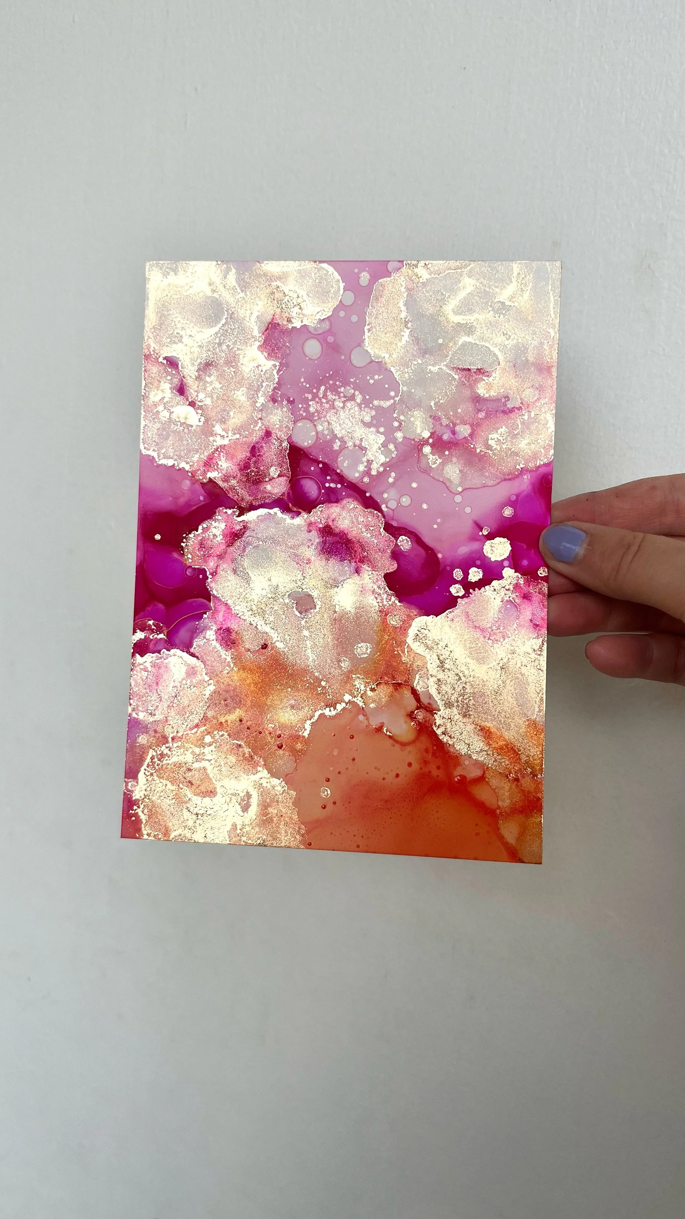 A hand holding a watercolor painting with pink, purple, orange, and silver accents on a plain white background. This is an aura painting and it comes with an aura reading. An aura reader & aura artist made this.