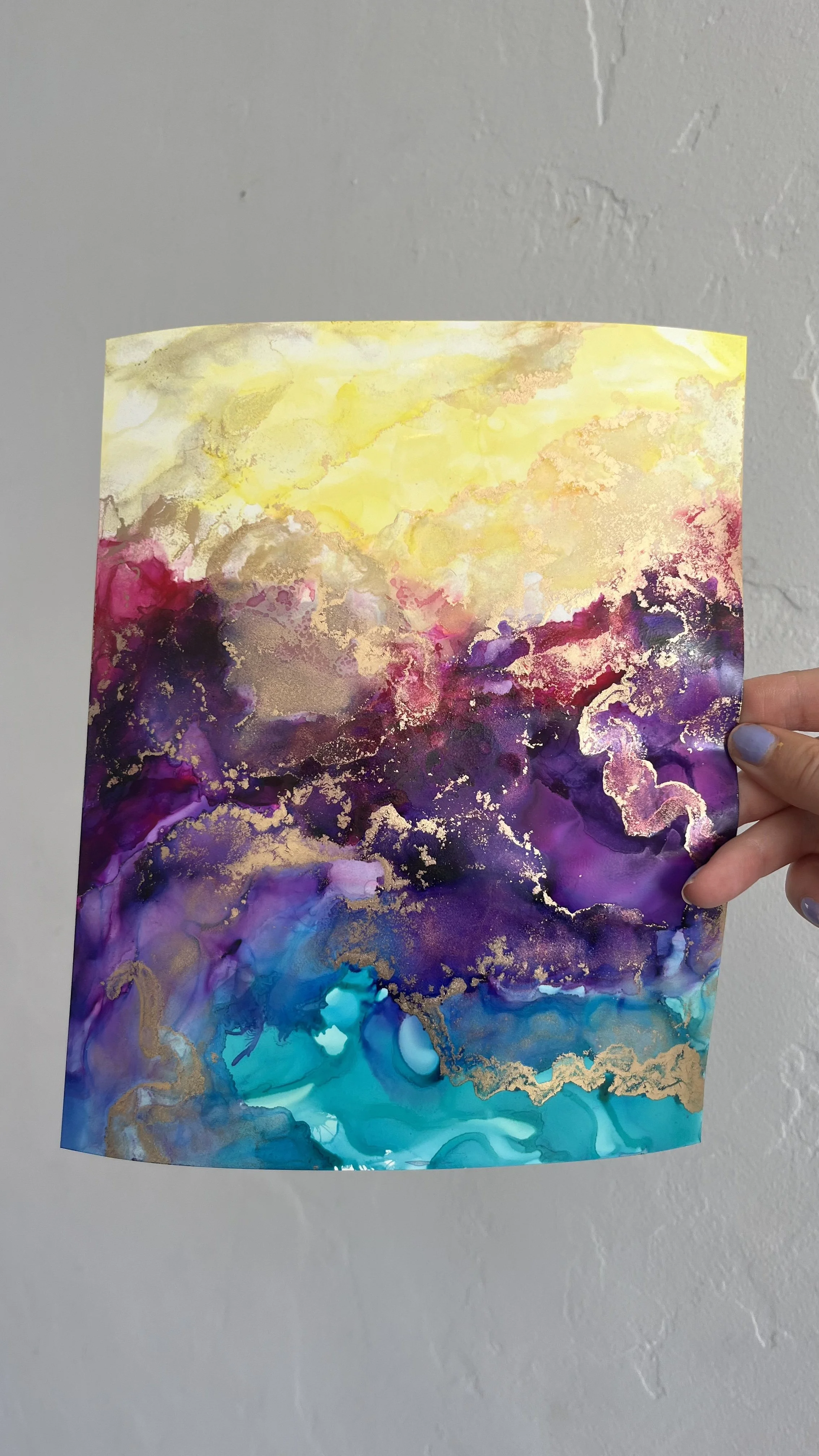 Hand holding a colorful abstract watercolor painting with shades of yellow, pink, purple, and blue, with gold accents, against a plain white wall. This is an aura painting and it comes with an aura reading. An aura reader & aura artist made this.