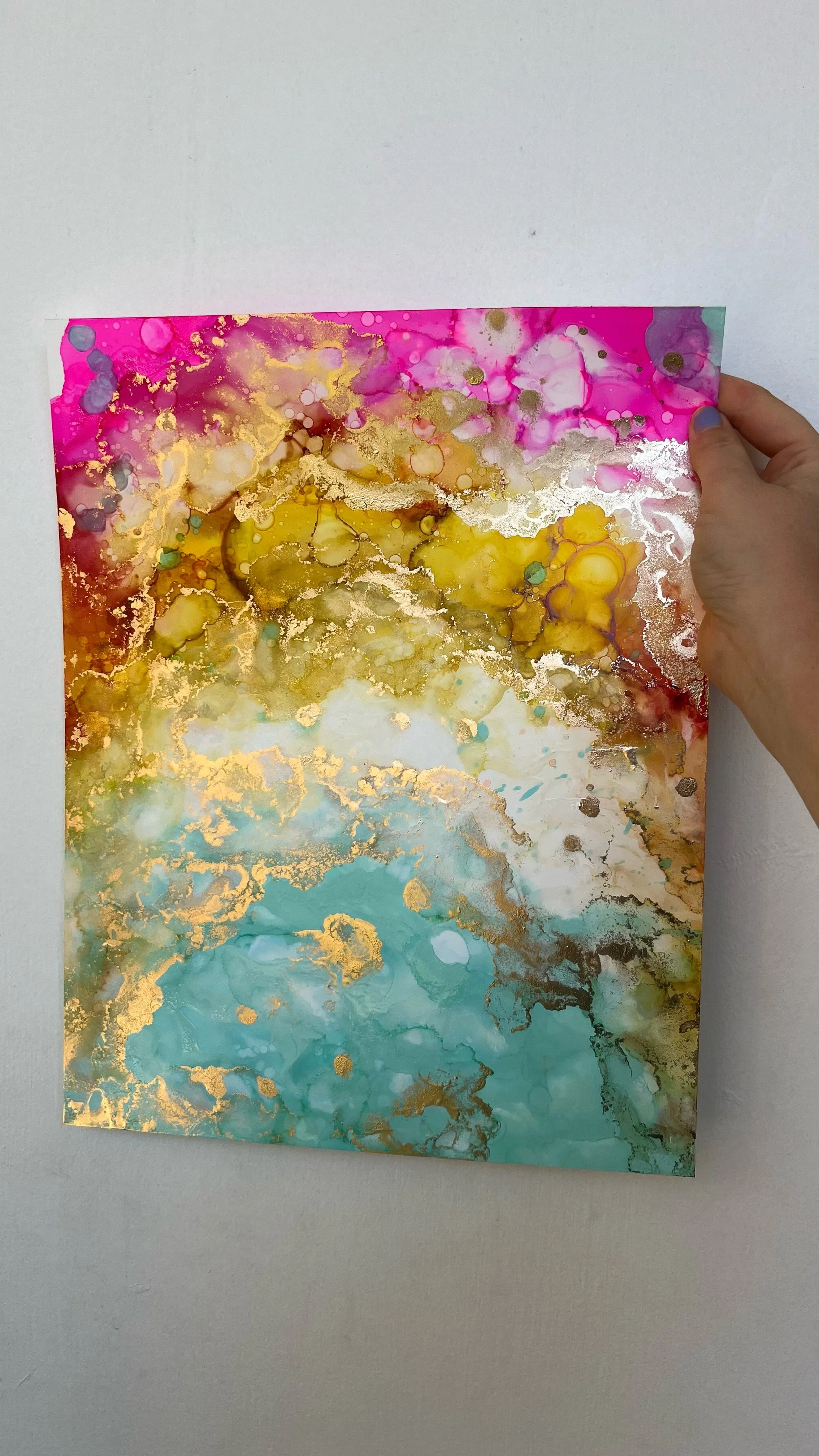 A hand holding a colorful abstract painting with pink, yellow, gold, and aqua hues and gold accents. This is an aura painting and it comes with an aura reading. An aura reader & aura artist made this.