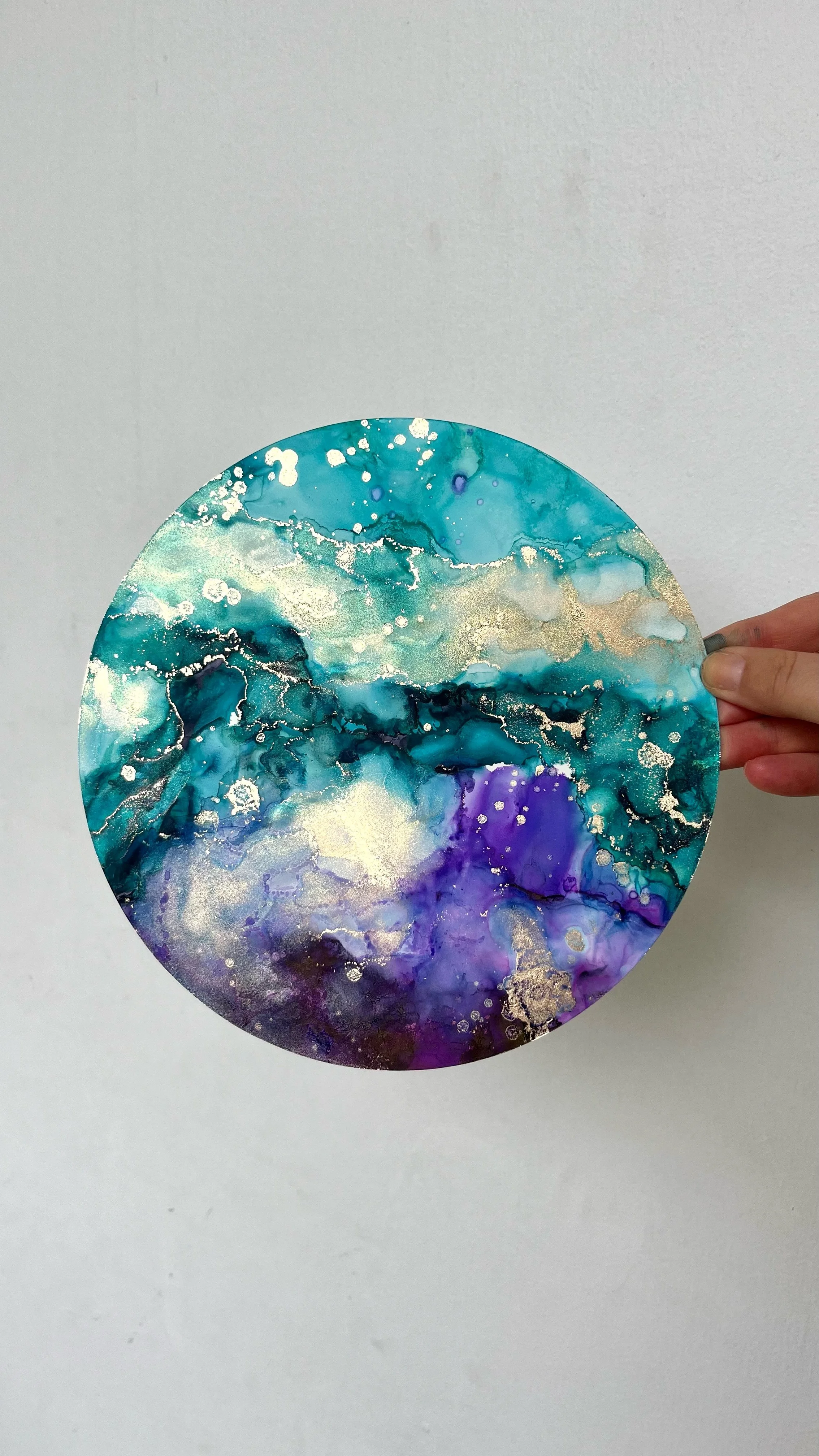 A circular abstract painting with blue, purple, white, and gold colors, held by a person's hand against a plain wall. This is an aura painting and it comes with an aura reading. An aura reader & aura artist made this.