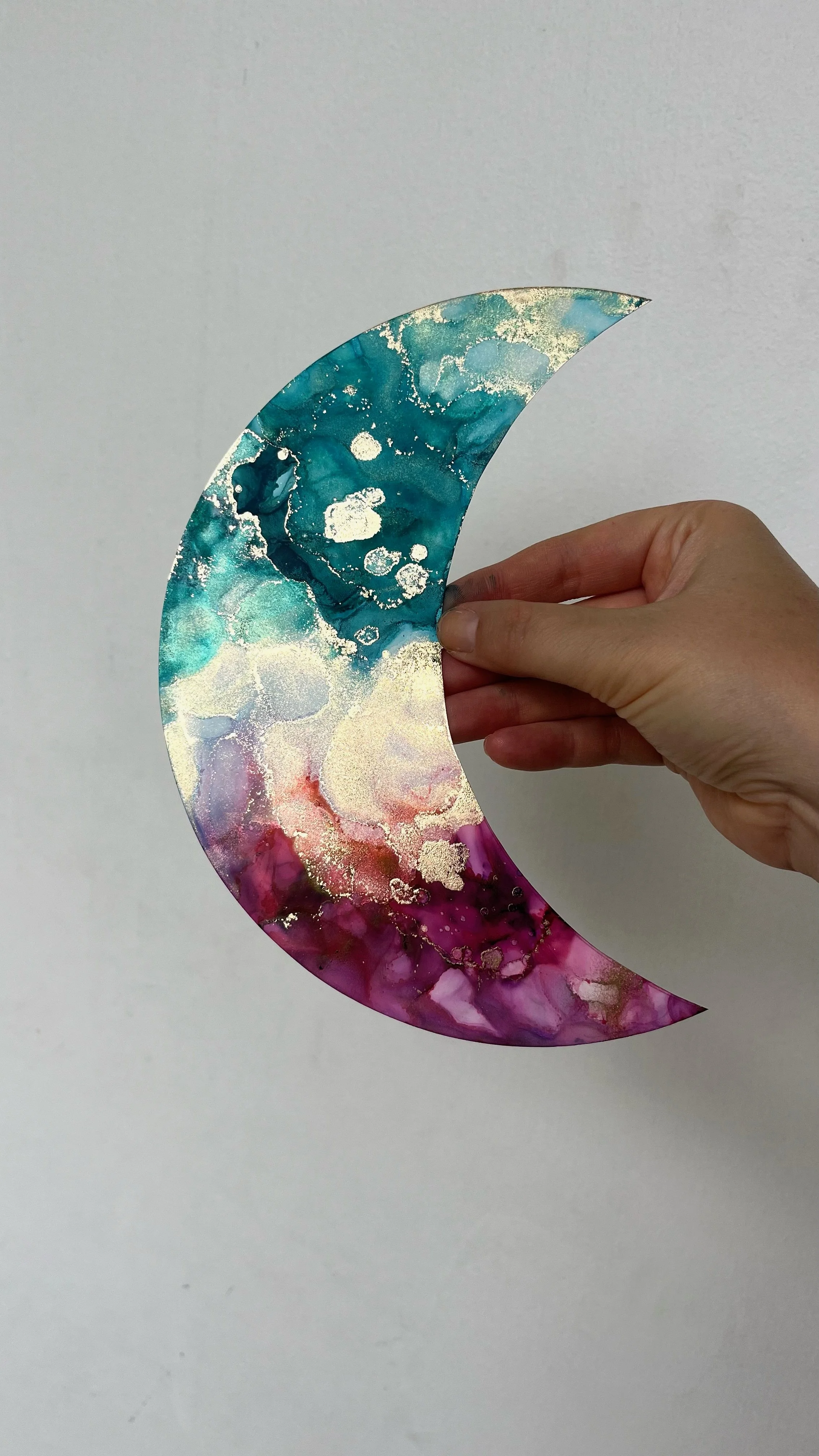 A hand holding a multicolored, abstract crescent-shaped piece resembling a moon, with a watercolor-like pattern in blue, white, pink, and purple, and gold accents. This is an aura painting and it comes with an aura reading. An aura reader & aura arti