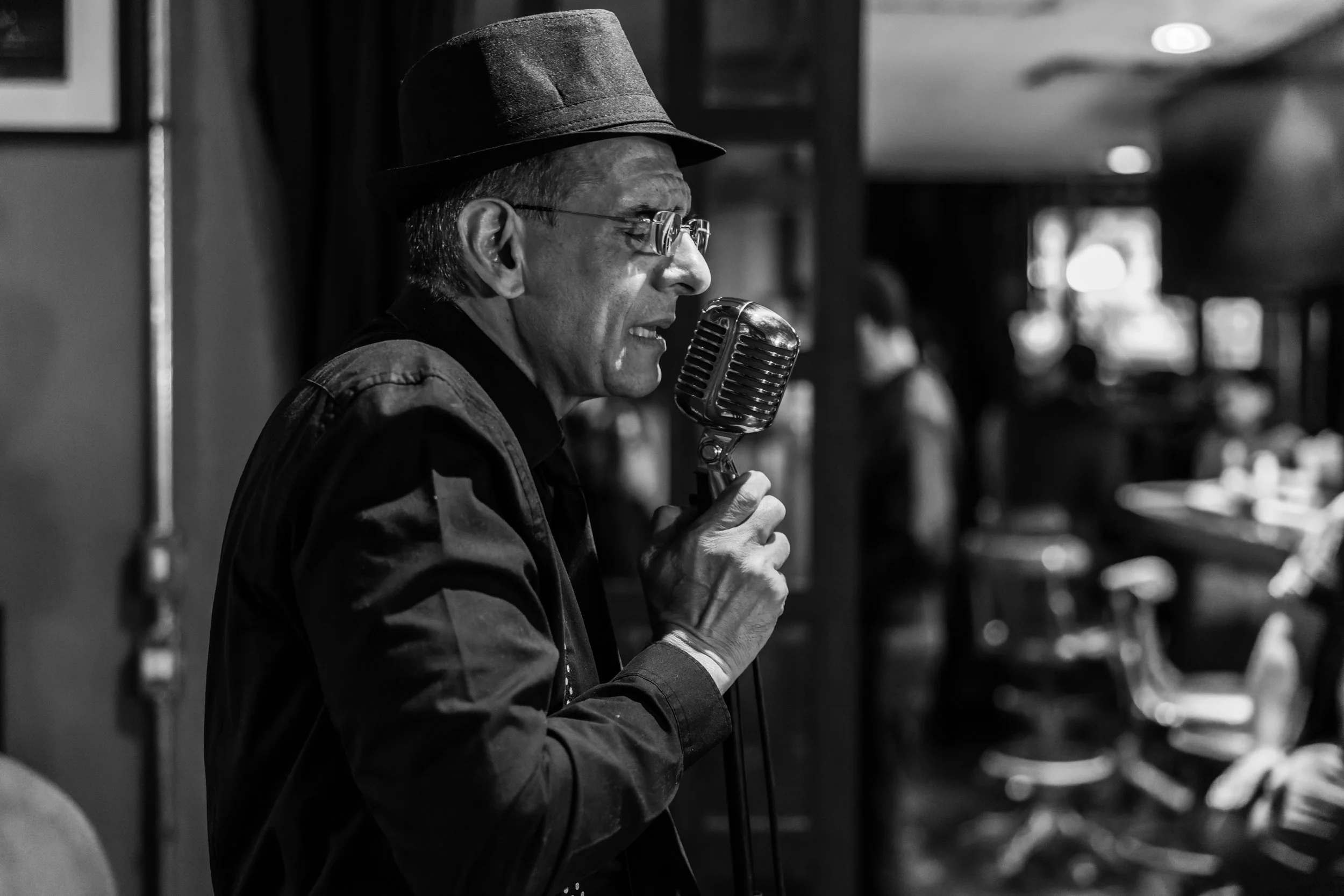 A black-and-white photo of a man wearing glasses and a hat, singing into a vintage microphone with a focused expression, in an indoor setting.