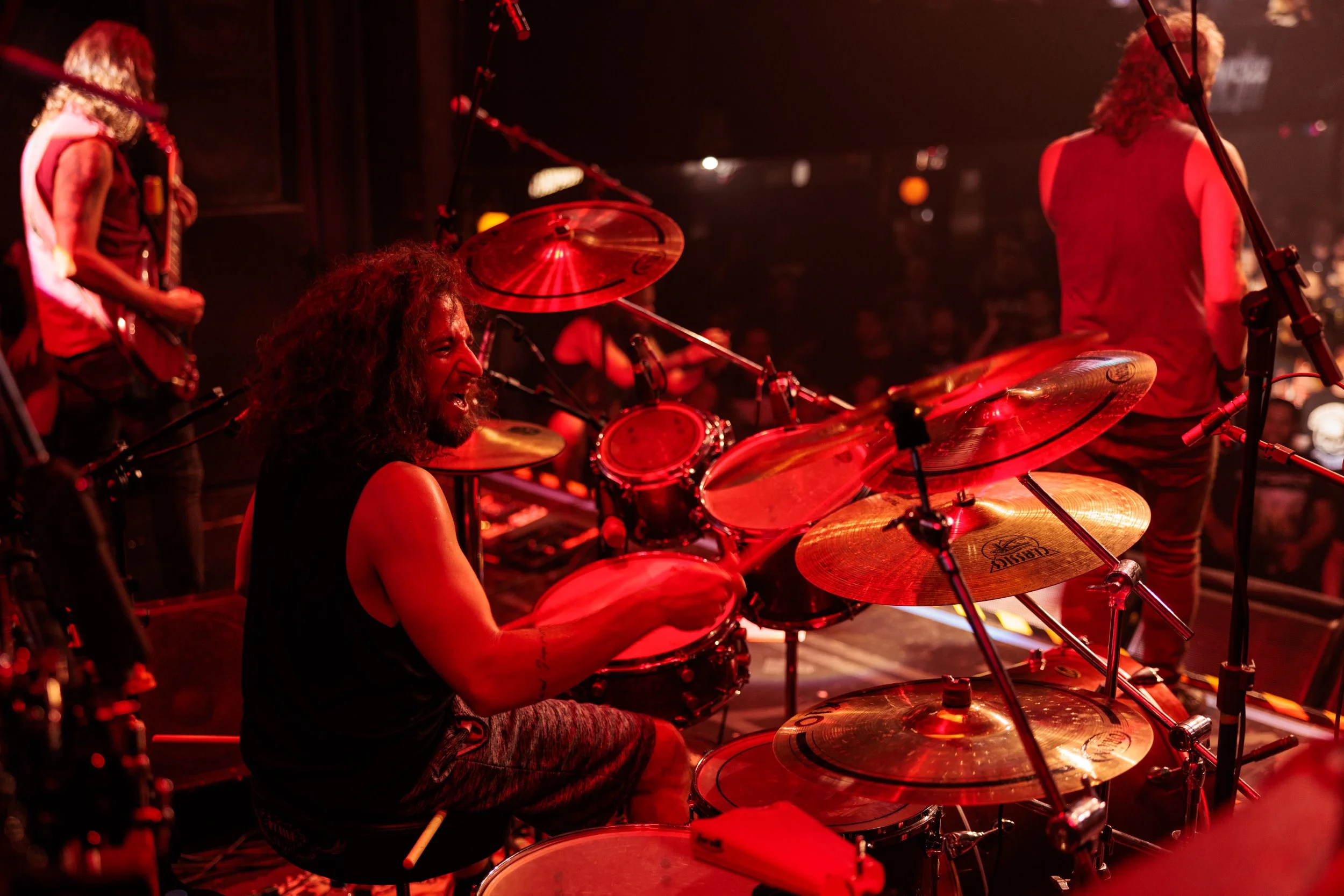 A drummer with curly hair playing a drum set on stage under red lights, with other musicians and microphones nearby.