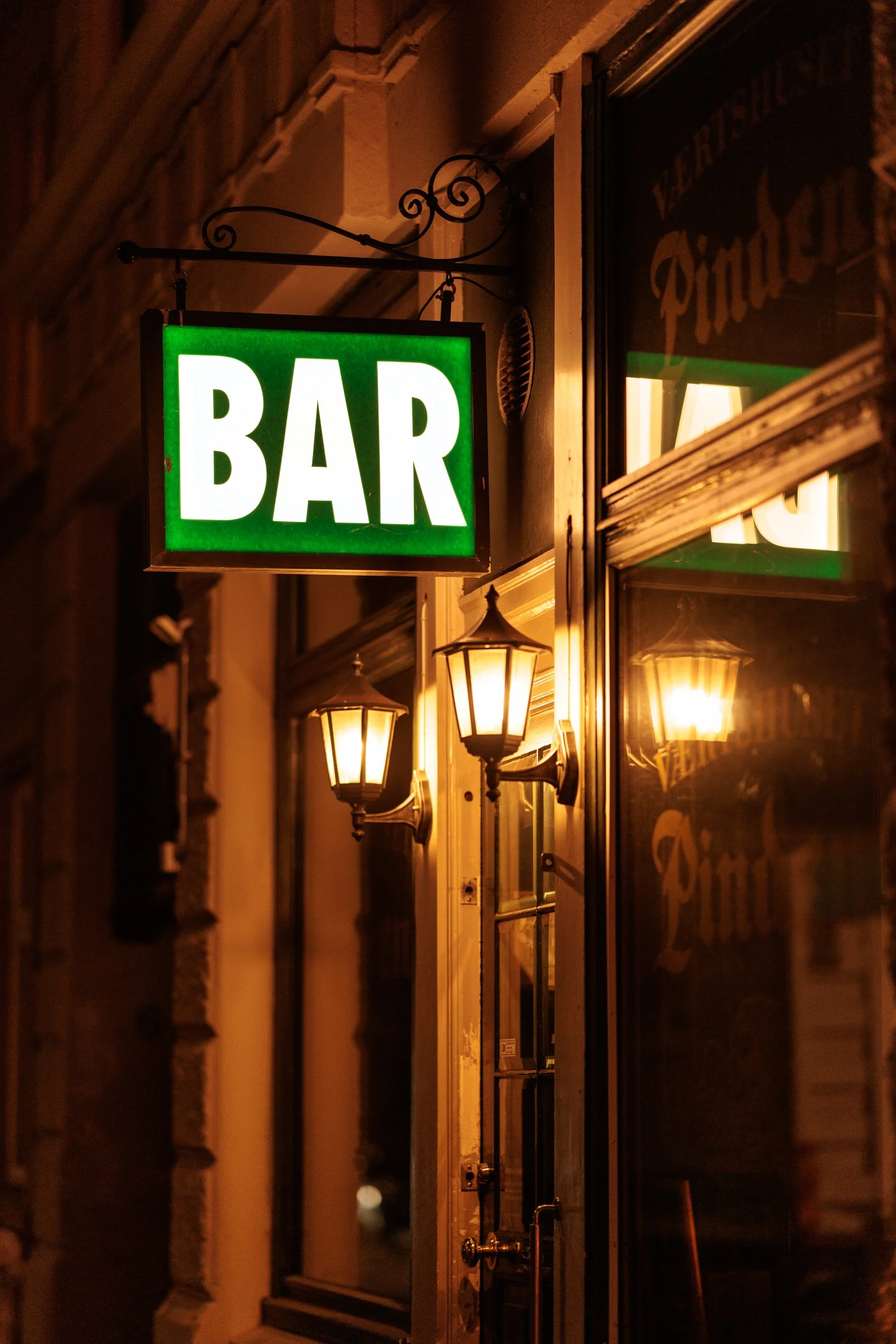A green illuminated sign with the word 'BAR' hanging outside a building at night. There are traditional-style wall-mounted lamps illuminating the exterior and a window reflecting part of the sign and lamps.