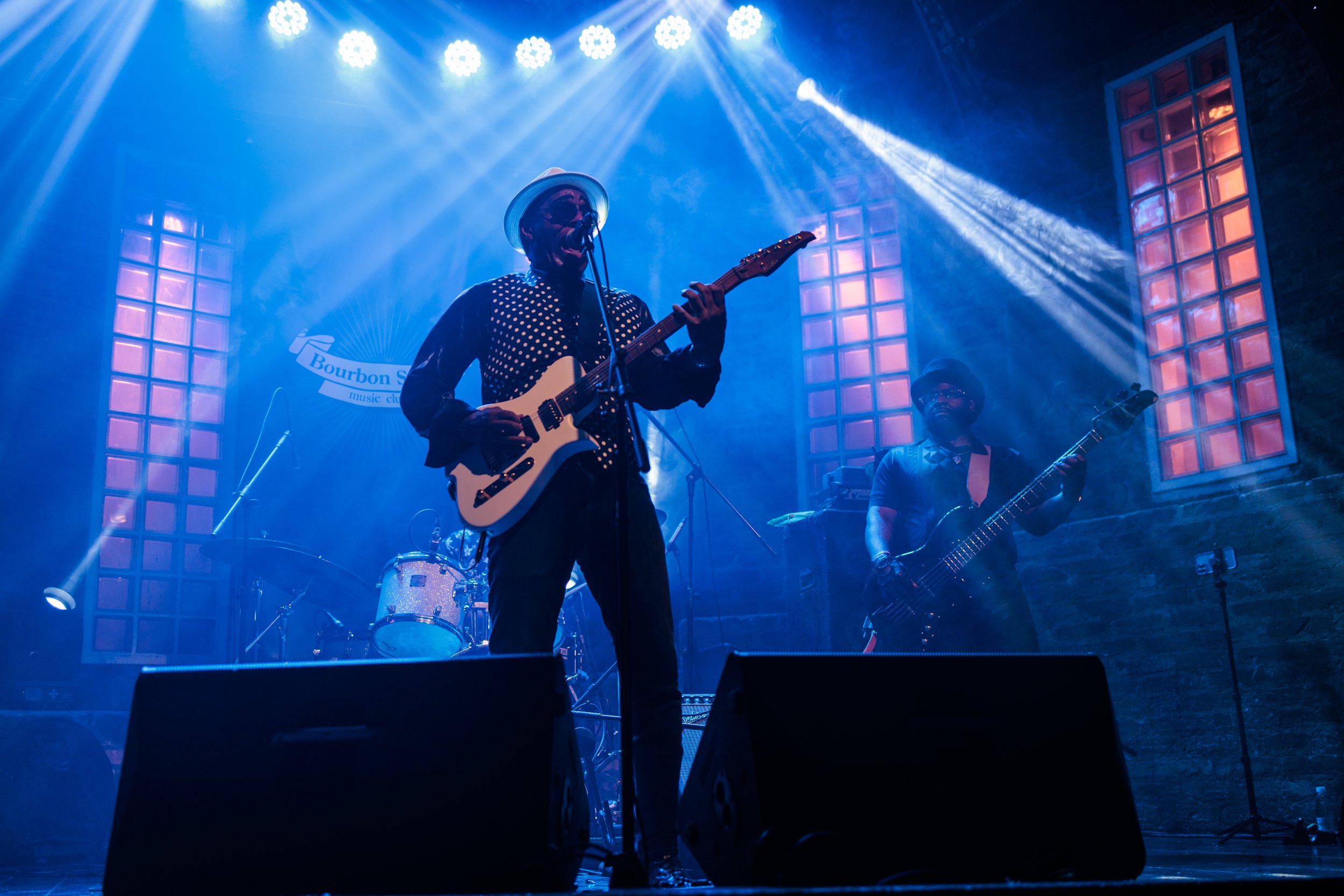 Musician wearing sunglasses and a hat playing electric guitar on stage with blue lighting and band members in the background.
