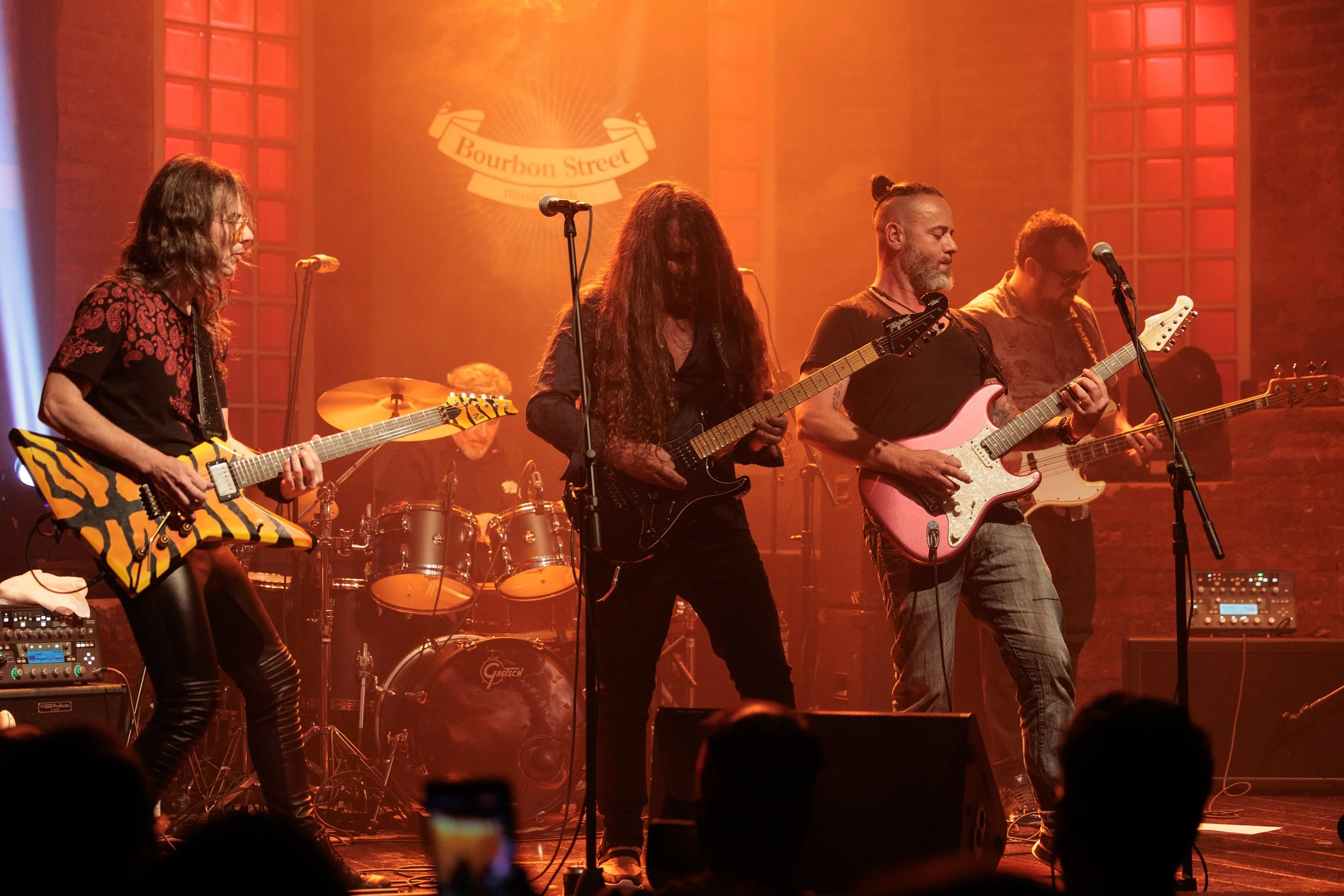 A live band performing on stage with guitarists, a drummer, and a bassist, under orange and red lighting with a Bourbon Street sign in the background.