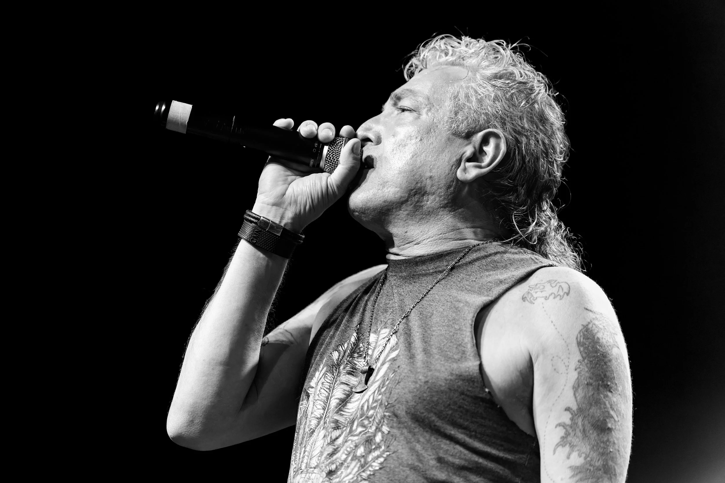 Black and white photo of a man with curly hair singing into a microphone, wearing a tank top and tattoos visible on his arm.