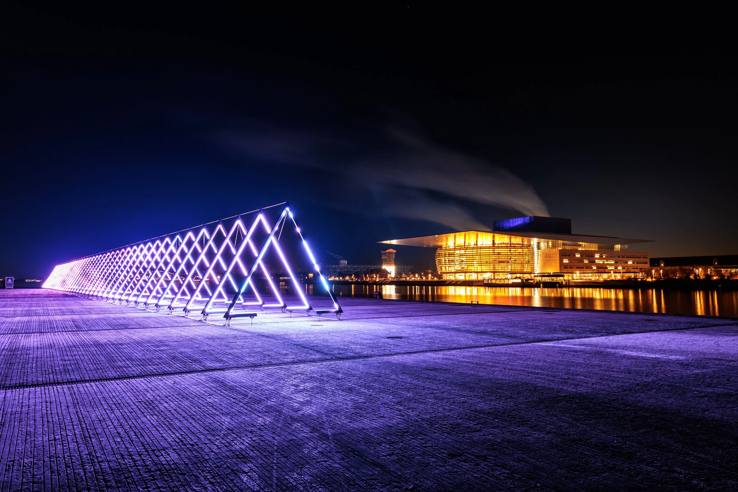 Night view of a waterfront with illuminated modern architectural structures, including a geometric purple-lit installation on the left and a large building with warm lighting on the right, reflecting in the water.