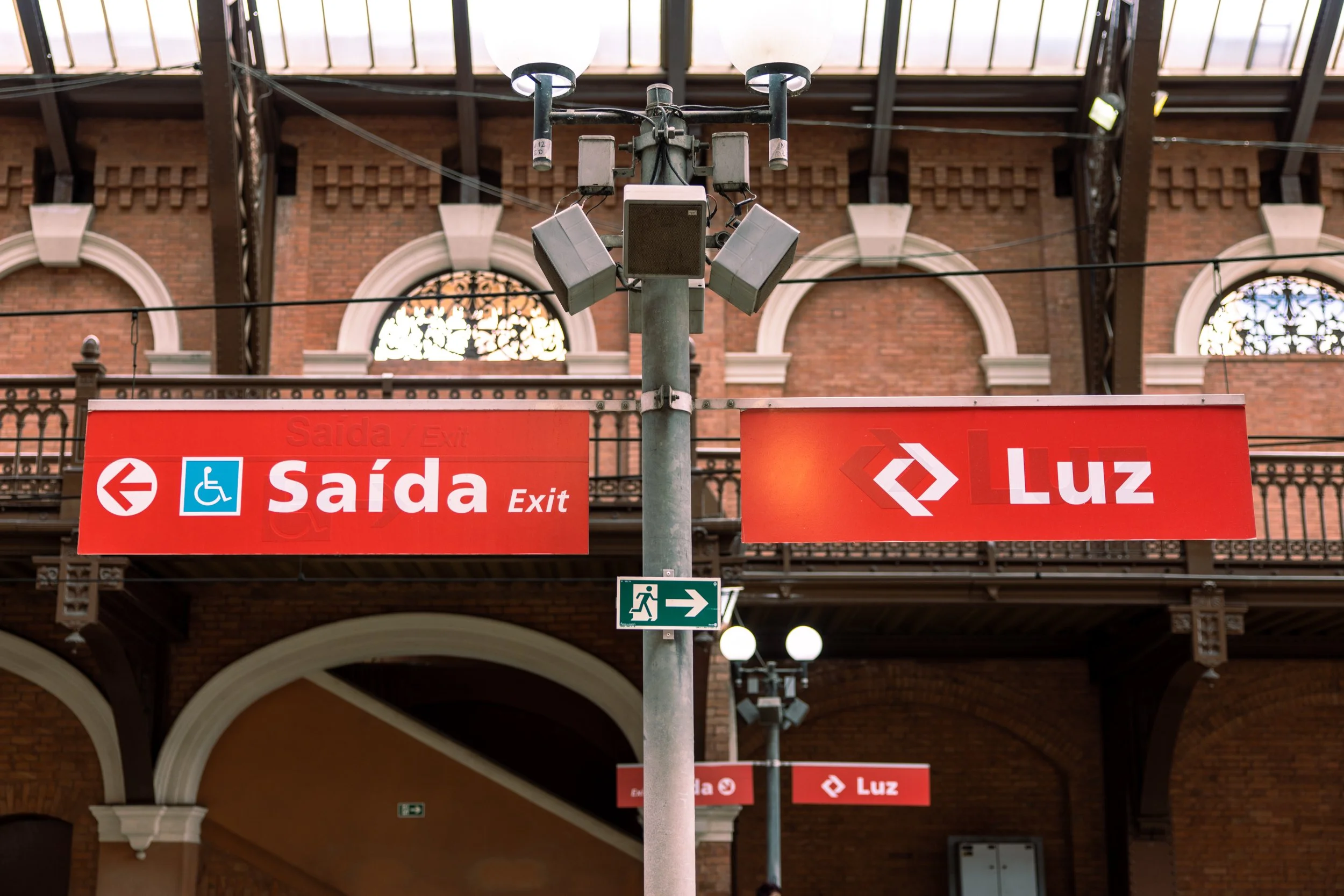 A tall pole with two large lamps on top, multiple speakers attached, and various signs in Portuguese. One sign points left with an icon of a wheelchair and the word 'Saída' (Exit), another points right with the word 'Luz' (Light), and a smaller green