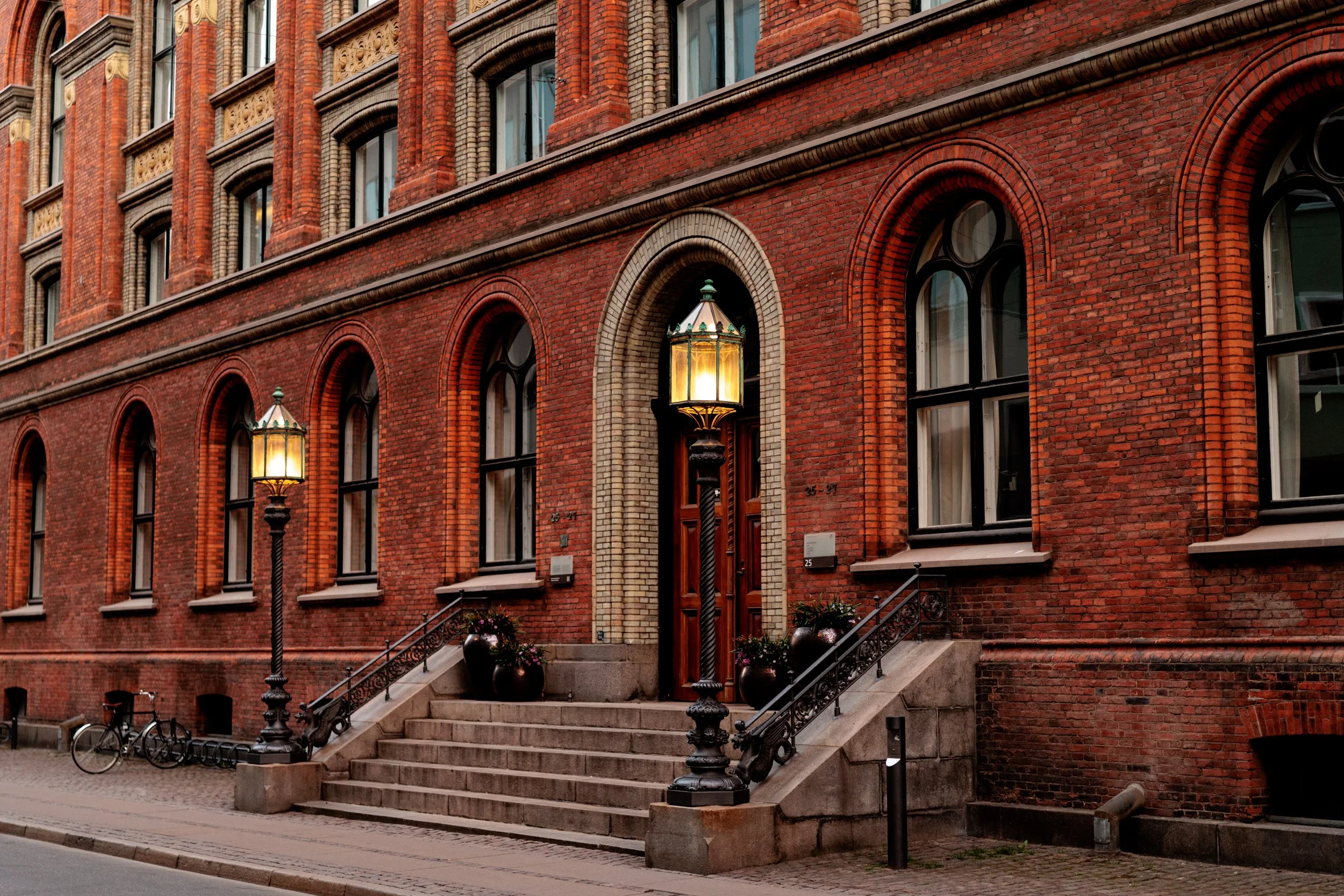 A brick building with arched windows and a set of stairs leading to the entrance, decorated with street lamps and potted plants.