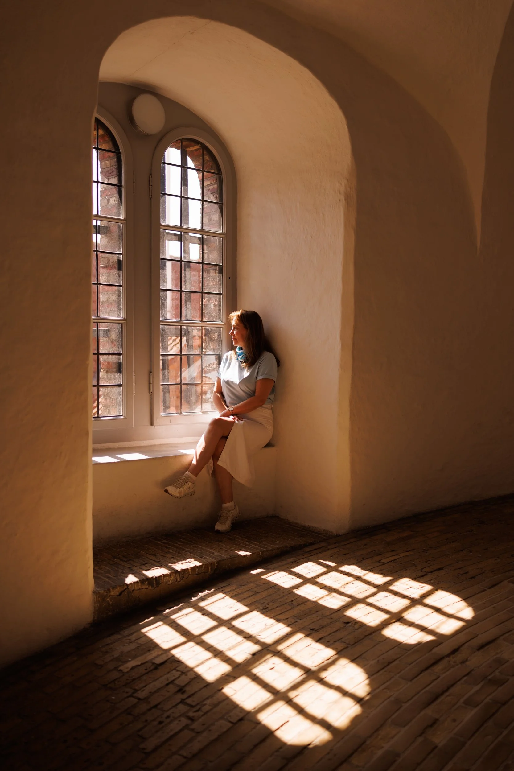 A woman sitting on a windowsill in a room with curved walls, sunlight streaming through a large, arched window, casting shadows on the brick floor.
