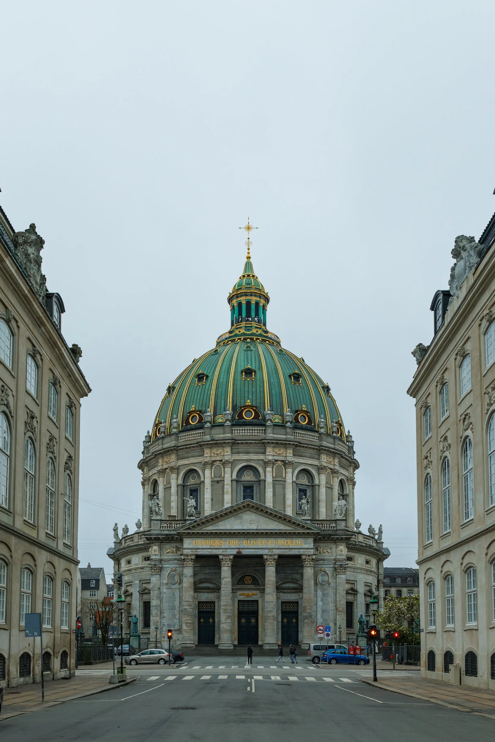 View of an ornate historic building with a domed green and gold roof, classical columns, and statues, seen from the street with pedestrians and cars, under an overcast sky.