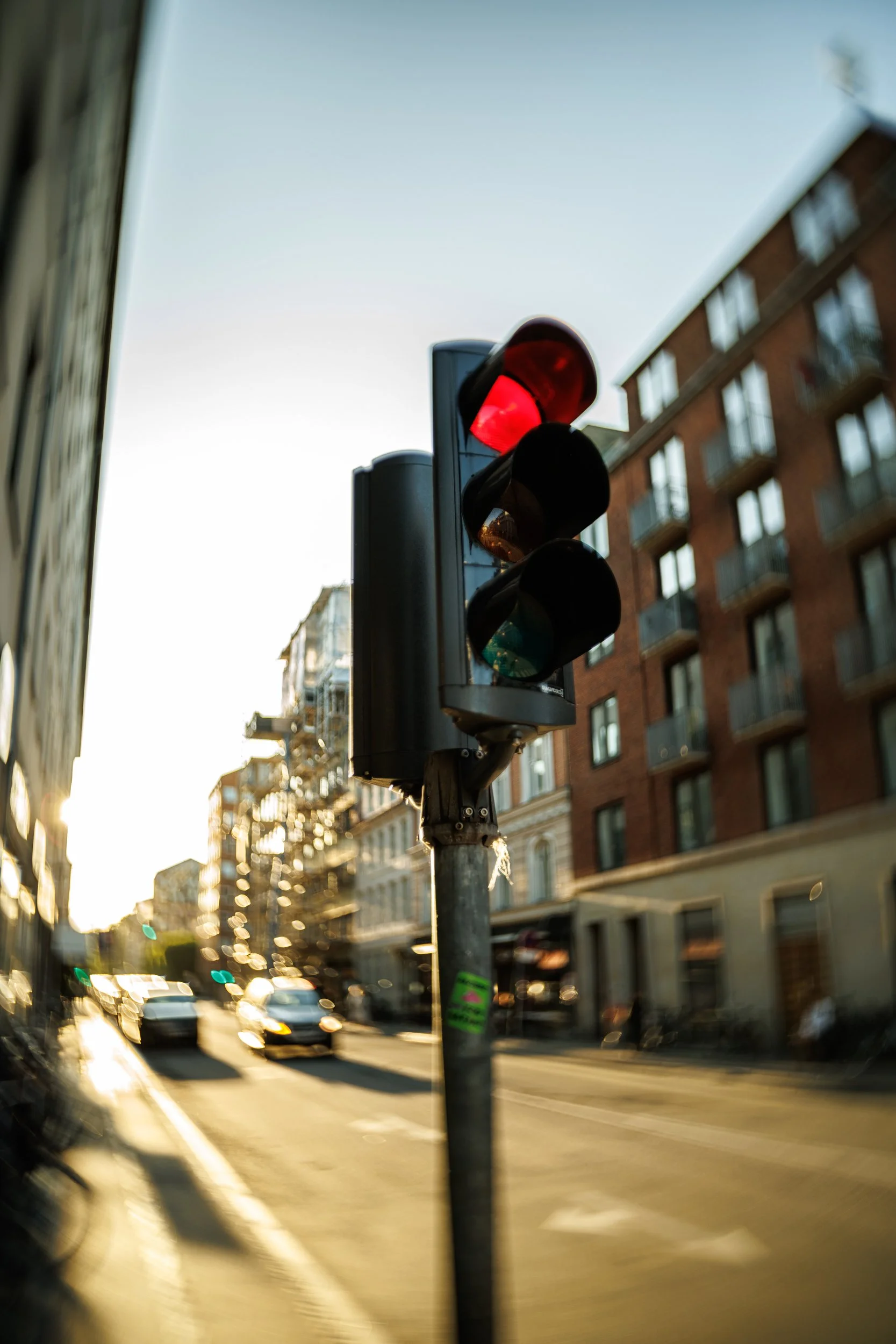 Traffic light with red signal in an urban city street scene during sunset, with buildings and cars on the road.