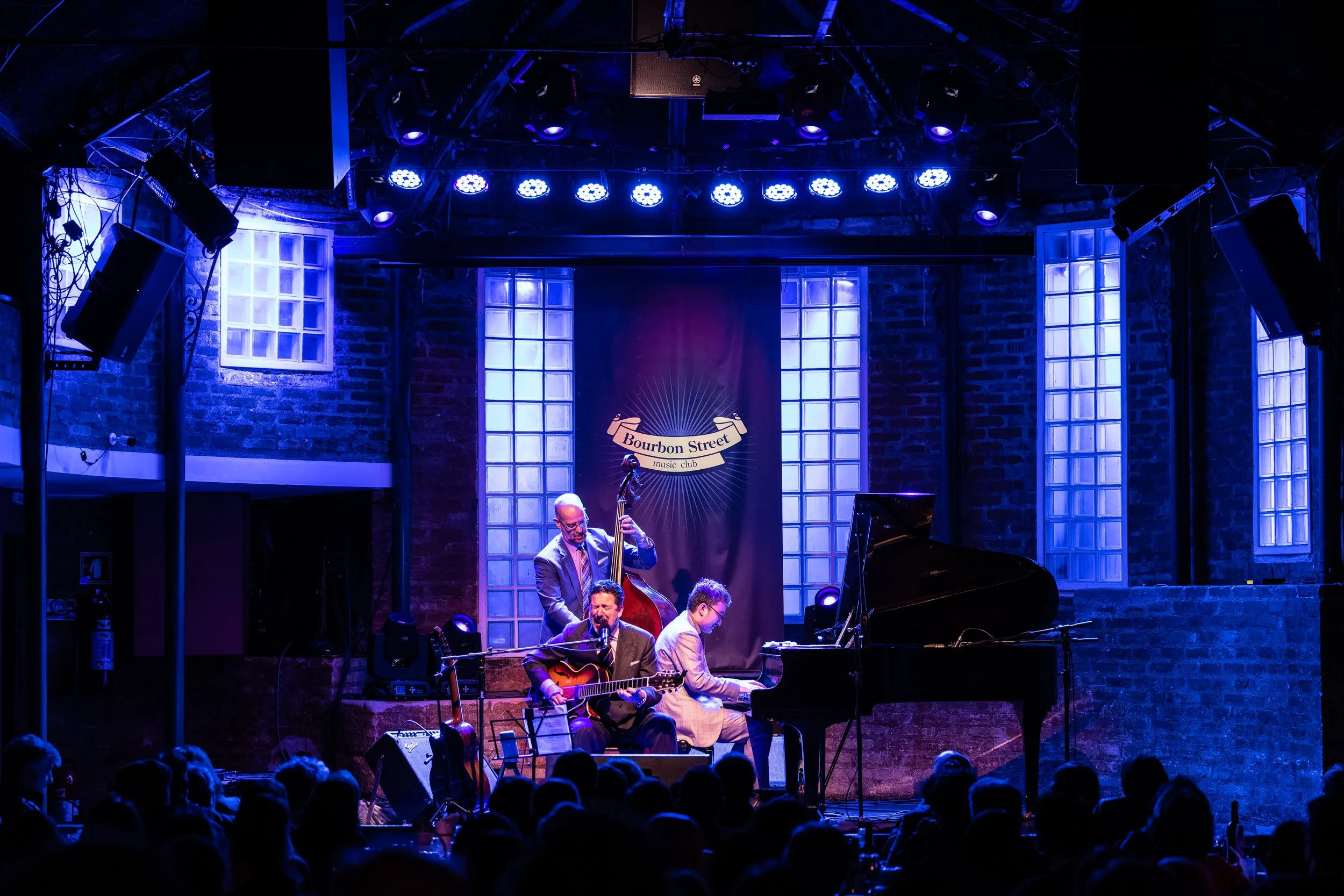 Live jazz band performing on stage at Bourbon Street music club, with blue stage lighting, brick wall backdrop, and audience in foreground.