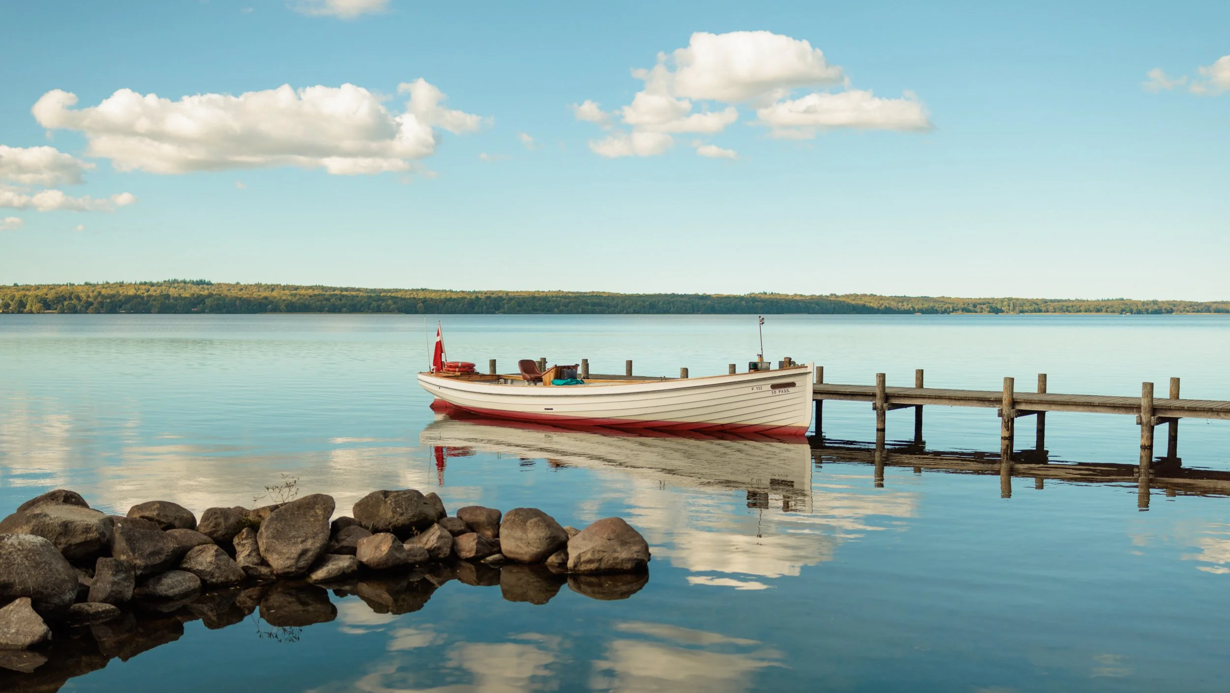 A white boat docked at a wooden pier on a calm lake with a clear blue sky and some clouds, rocky shoreline in the foreground, and trees in the distant background.