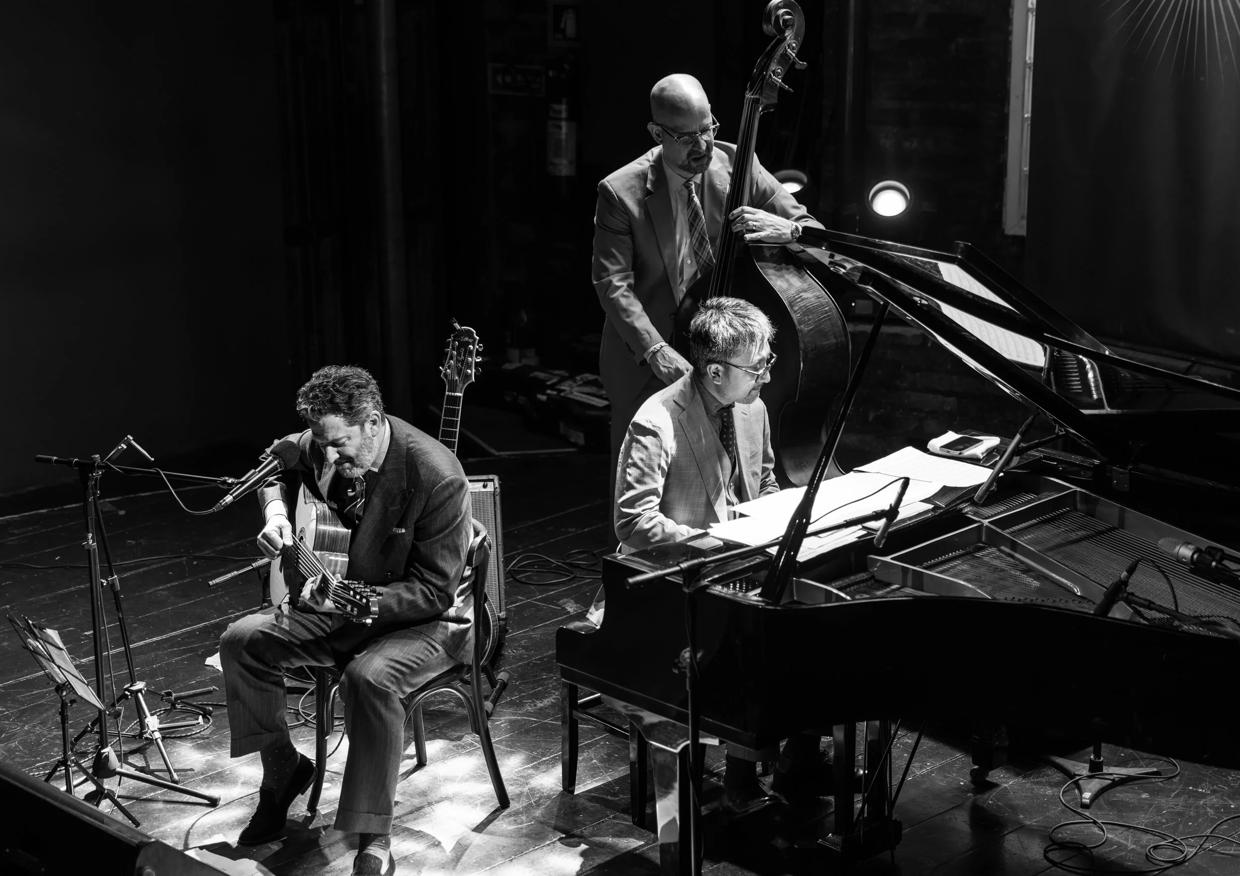Black and white photo of four musicians performing on stage. One is playing an acoustic guitar and singing into a microphone, another is playing a double bass, and two are at a piano, one of them reading sheet music. The stage is dimly lit with spotlights.