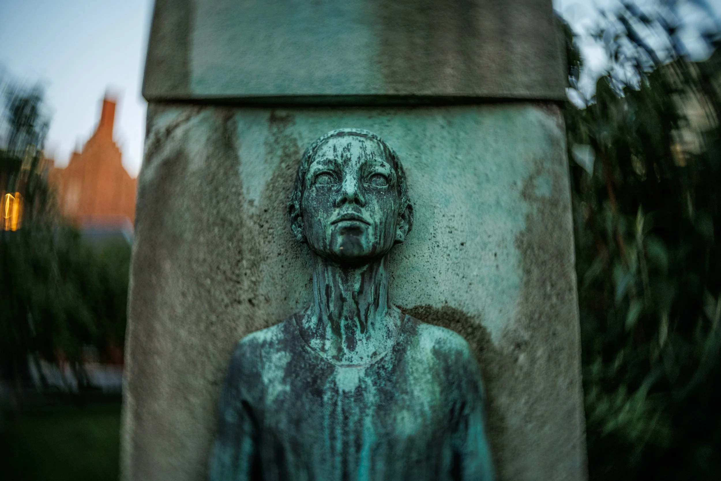 A bronze bust of a woman with long hair, mounted on a stone pillar in an outdoor setting.