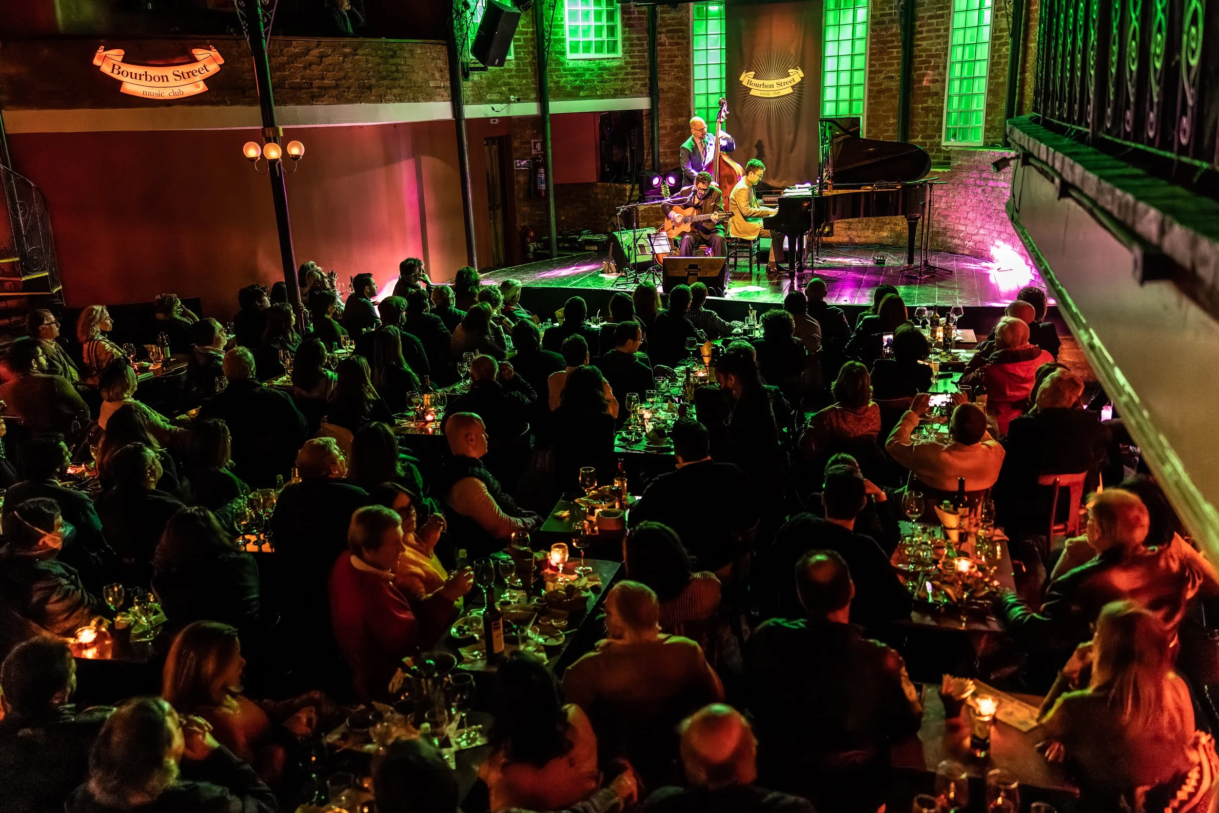 A live music performance at Bourbon Street music club, with a band playing on stage and an audience seated at tables, enjoying drinks in a dimly lit room with green and pink lighting.