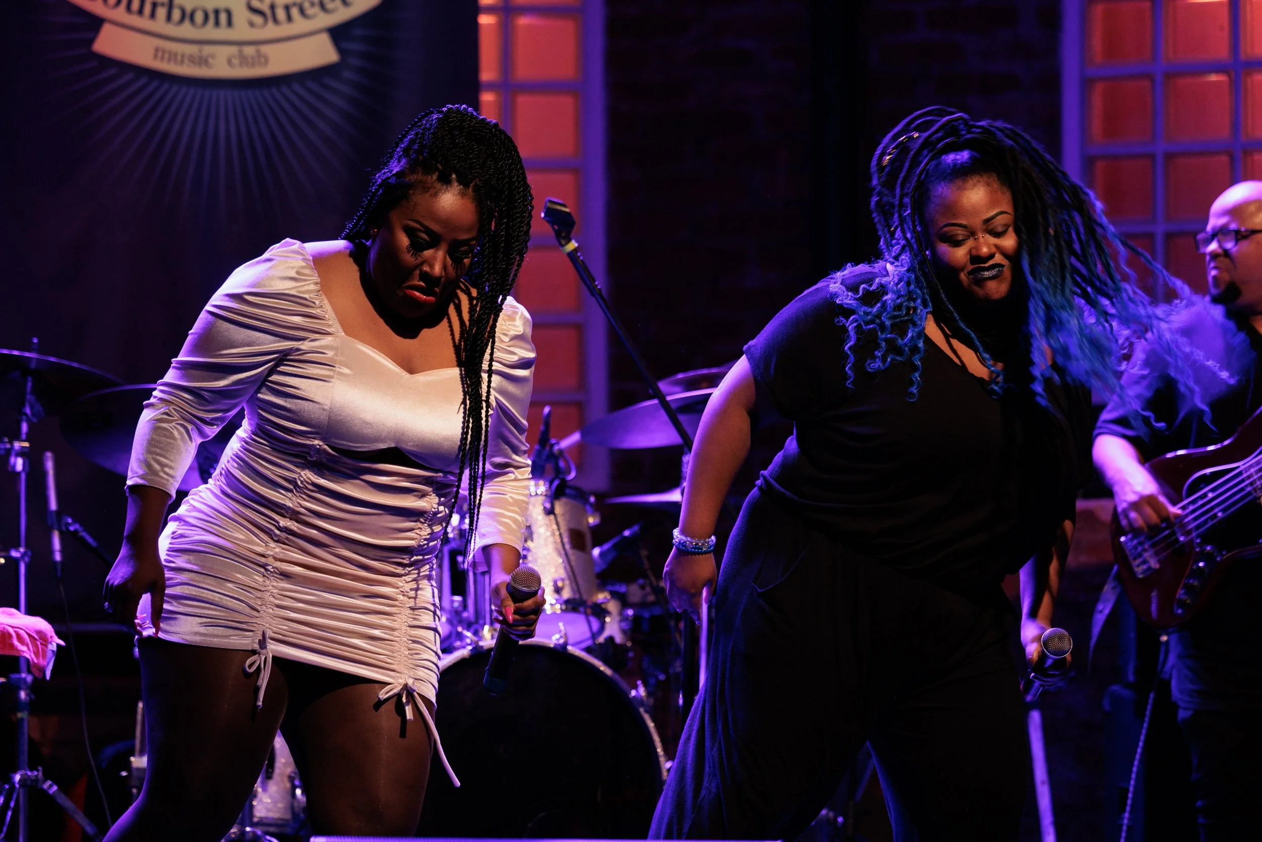 Two women standing on stage at a music club, possibly about to perform, with musical instruments and a drummer in the background. One woman holds a microphone, the other has blue-highlighted dreadlocks.