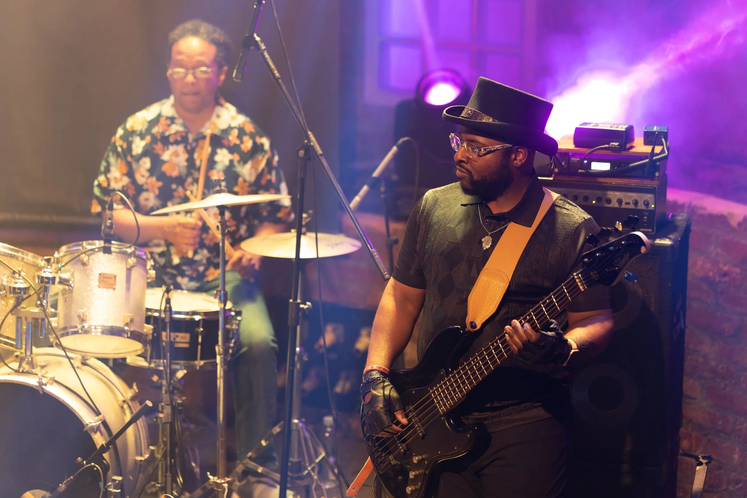 A musician playing an electric guitar in a dimly lit room with purple and pink stage lights, another person in the background sitting behind a drum set with a floral shirt and glasses.
