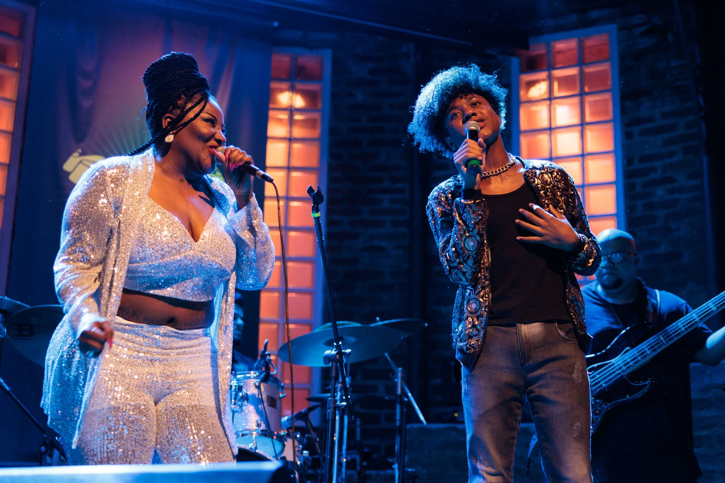Two singers performing on stage with microphone stands, one wearing a shiny silver outfit with braids in her hair, the other with curly blue hair and a patterned jacket, in a dimly lit venue with brick walls and orange-lit windows in the background.