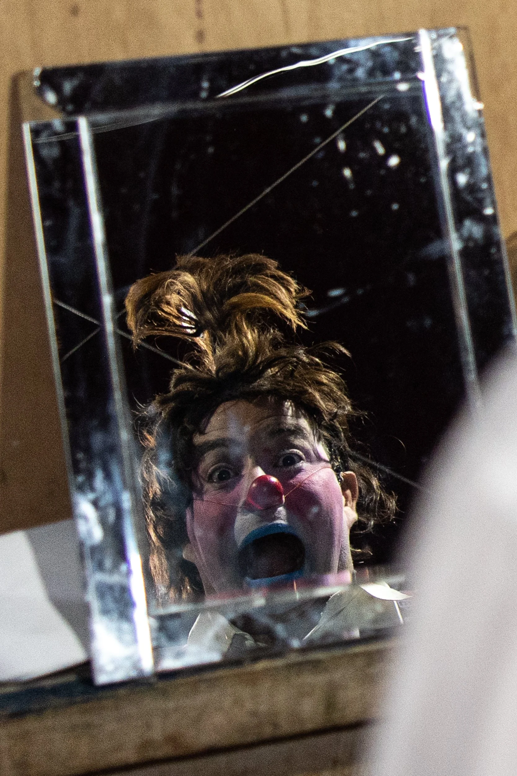 A reflection of a person with curly hair, wearing clown makeup and a red clown nose, is seen in a mirror on top of a wooden surface. The person's mouth is wide open, expressing surprise or excitement.