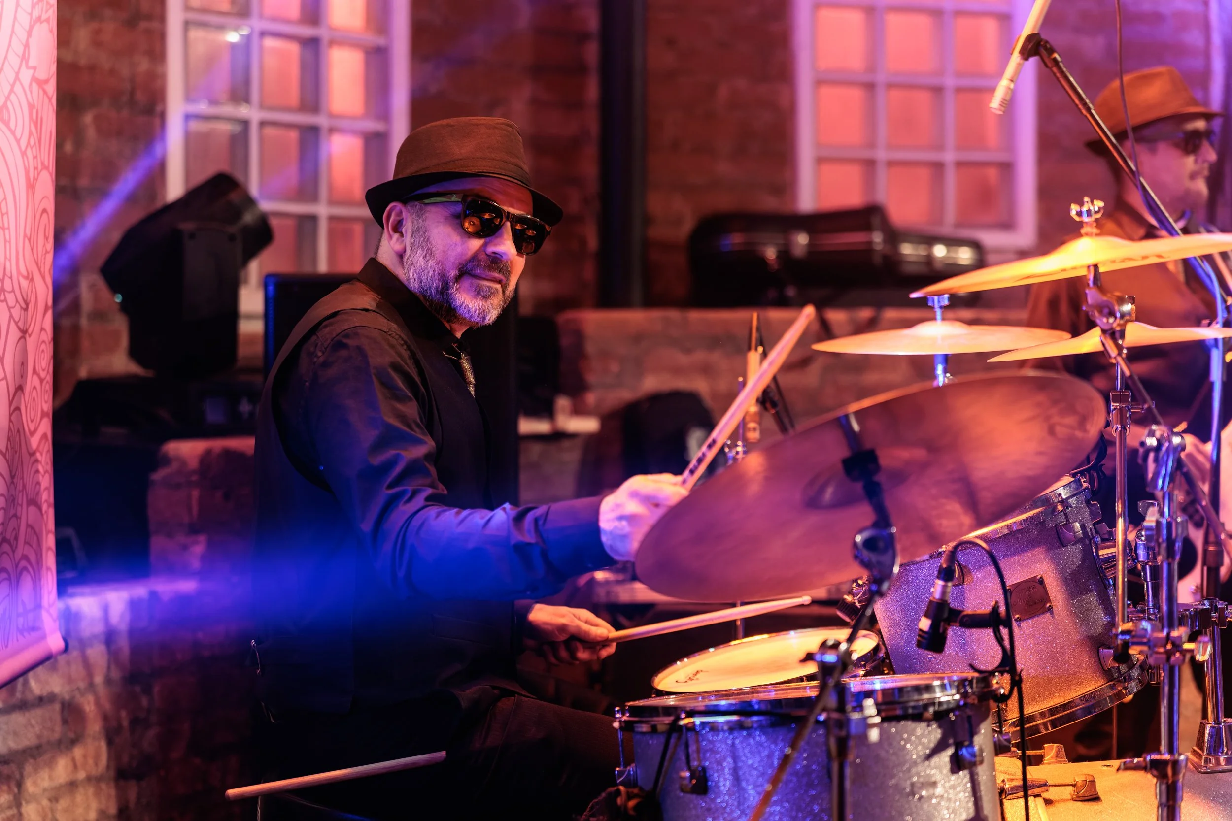A man wearing sunglasses and a brown hat playing drums in a dimly lit indoor setting with warm lighting and brick walls.