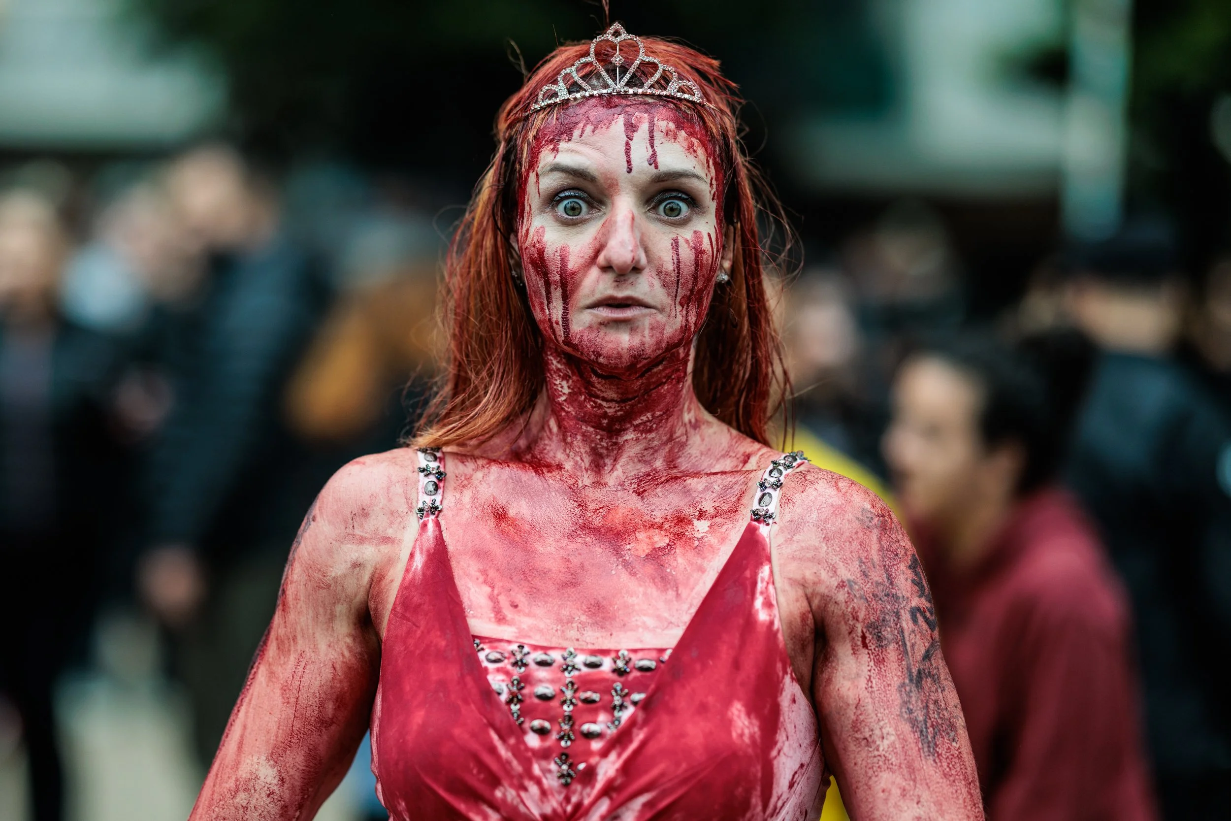 A woman with red hair and wide eyes, wearing a crown and a red dress, covered in red paint or blood, standing outdoors in a crowded area.