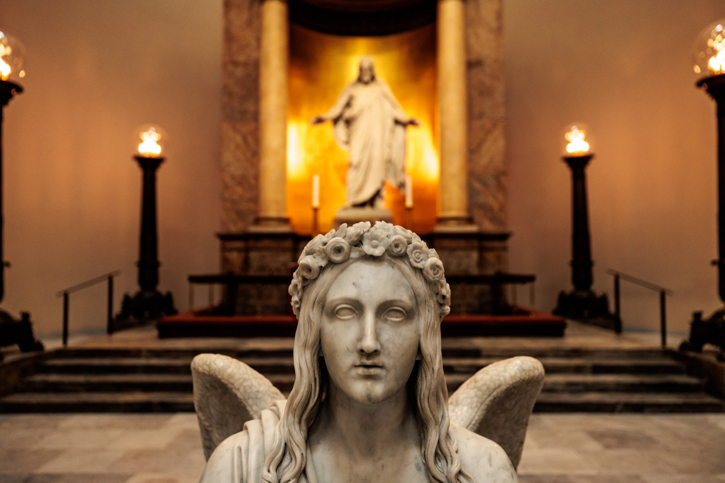 Marble sculpture of a woman with long hair and a floral crown, with snakes at her arms, in front of a religious altar with a statue of Jesus Christ with open arms, lit candles, and steps leading up to it.