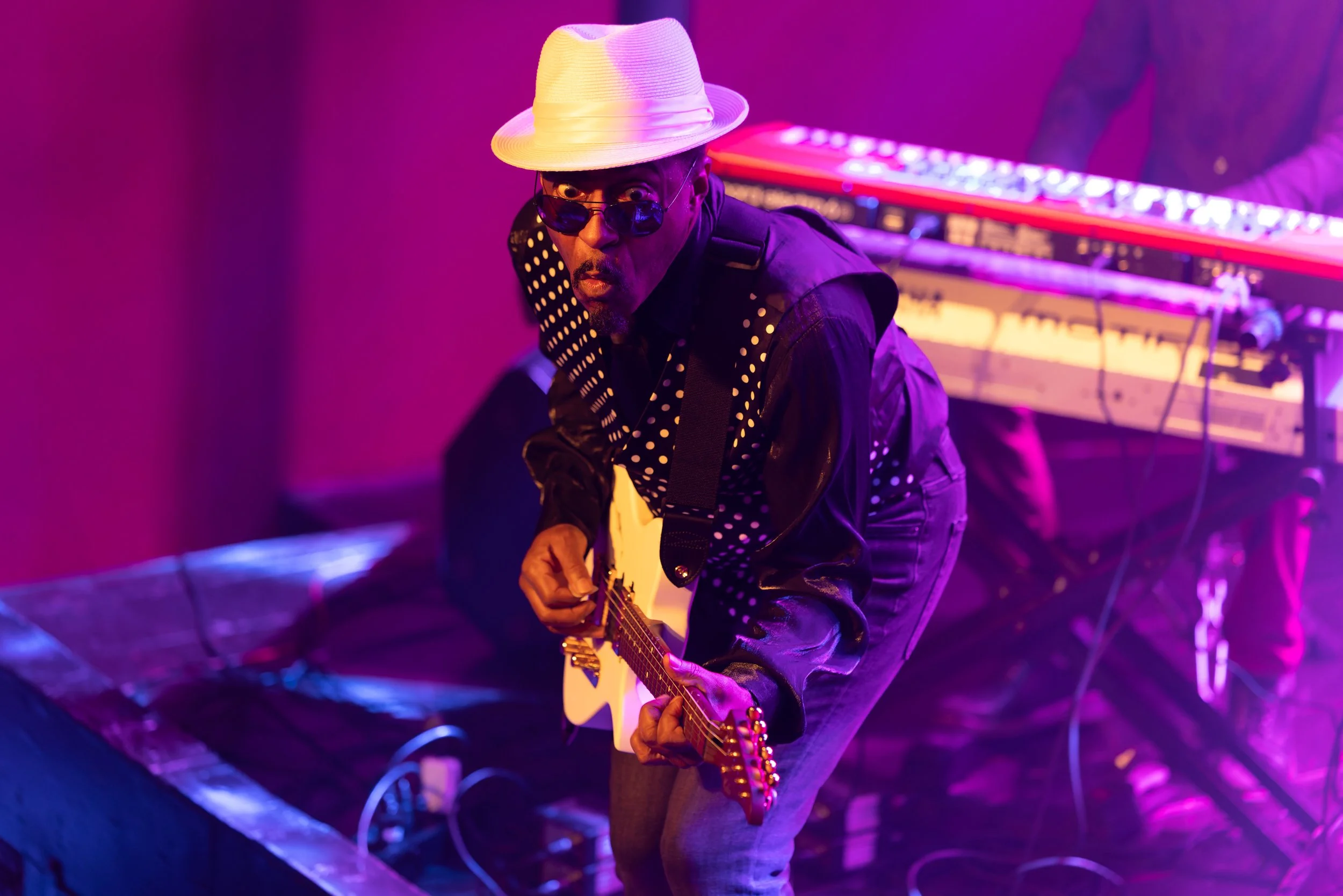 A man wearing a white fedora hat and sunglasses, playing an electric guitar on stage with purple and pink lighting.