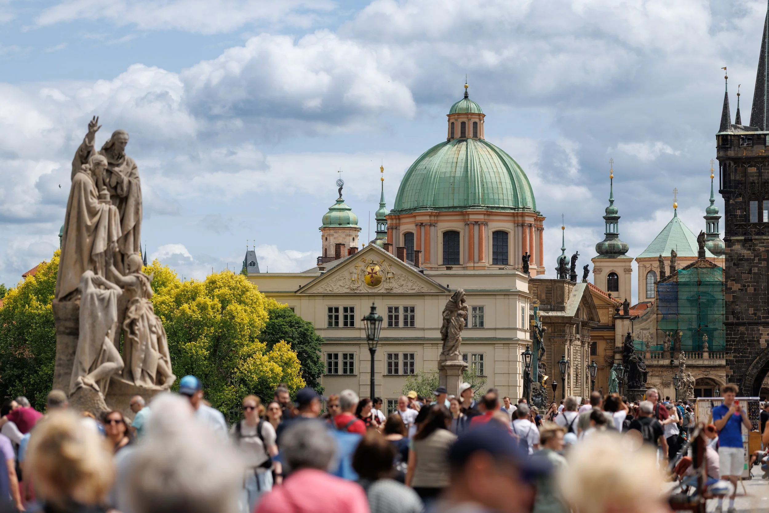 A crowded outdoor scene with many people walking and gathering in a historic European city square, featuring classical architecture, statues, and a large building with a green dome under a partly cloudy sky.