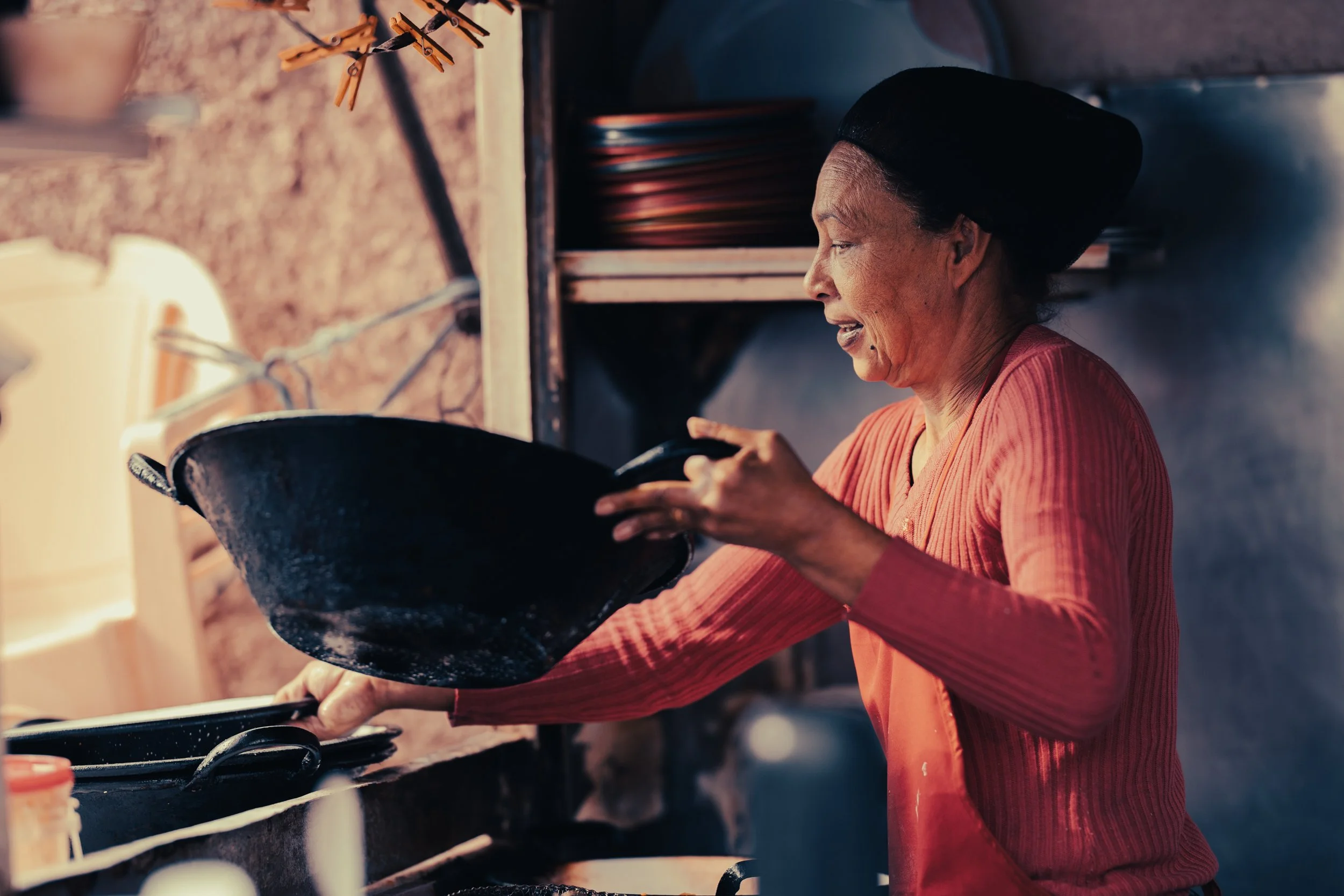 An older woman with a black headscarf cooking on a stove, holding a black frying pan and smiling.