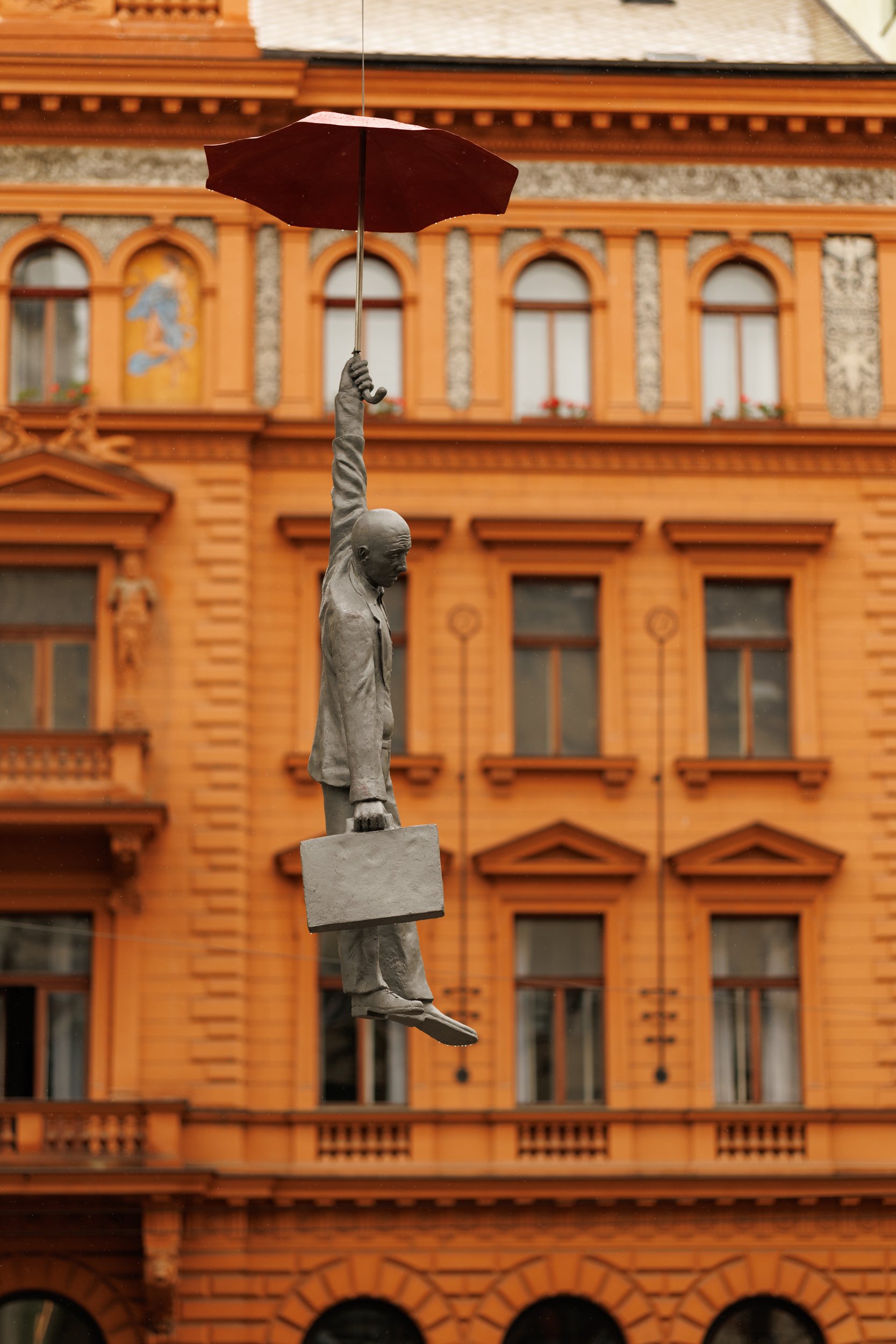 A street art sculpture of a man hanging from a street lamp, holding an umbrella, with a decorative orange building in the background.