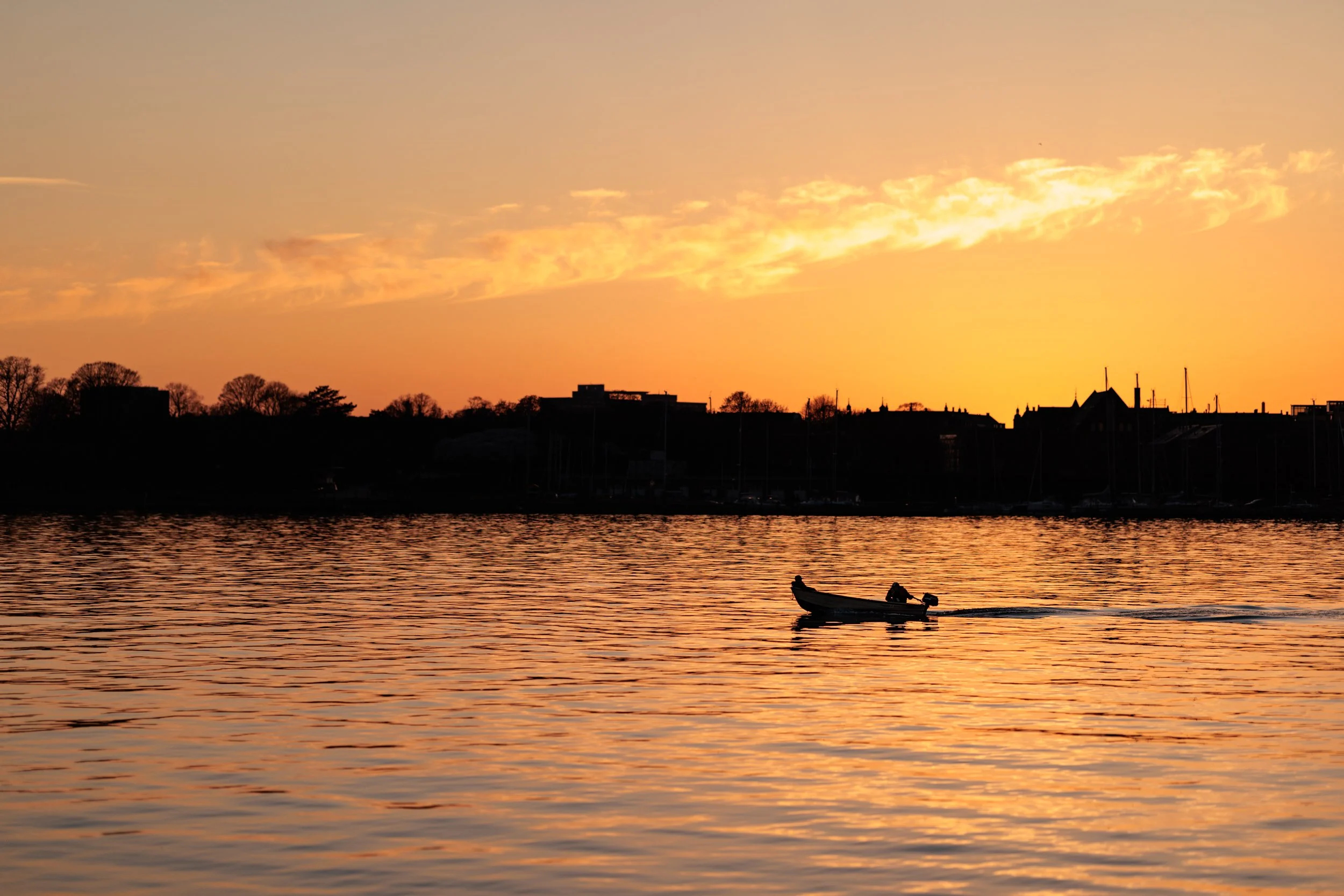 A sunset over a body of water with a small boat carrying a person, and a distant shoreline with trees and buildings.