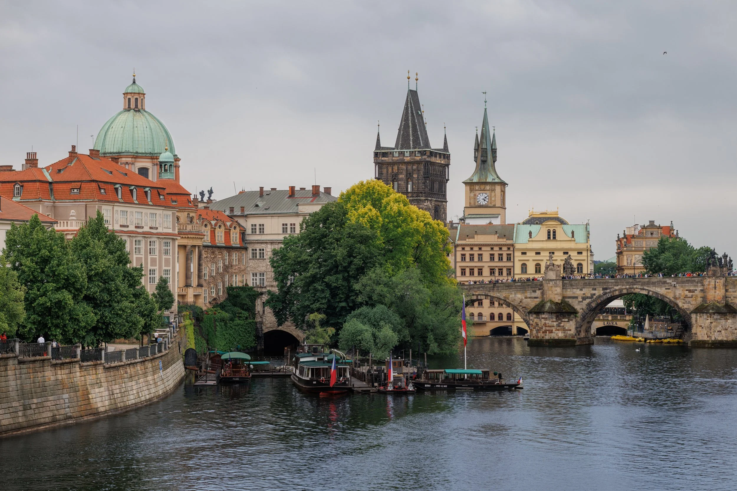 View of historic buildings including a domed church with a green roof, towers with spires, and a bridge over a river with boats and trees along the riverbank under a cloudy sky.