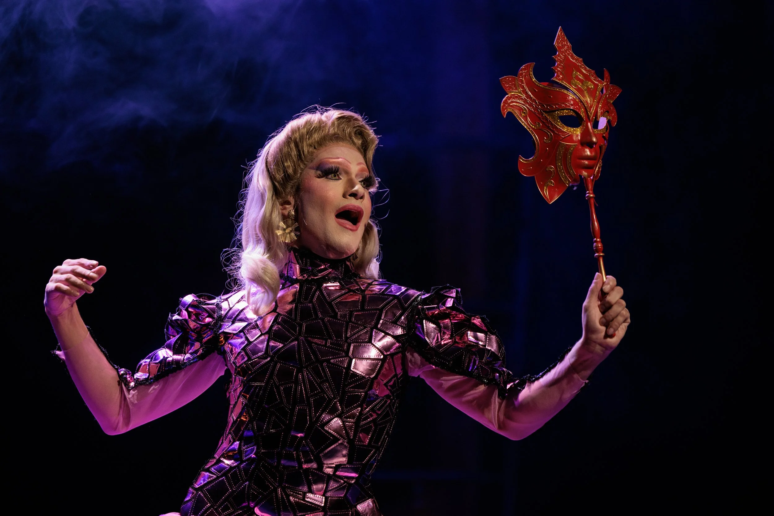 Performer in colorful costume holding a decorative mask on a stick, performing on stage against dark background.